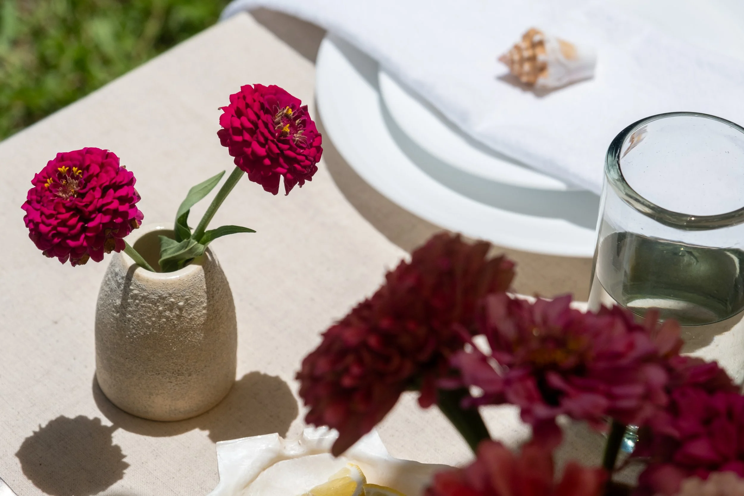 A beige textured vase holding two pink flowers on a table outdoors with a white plate and glass of water nearby.