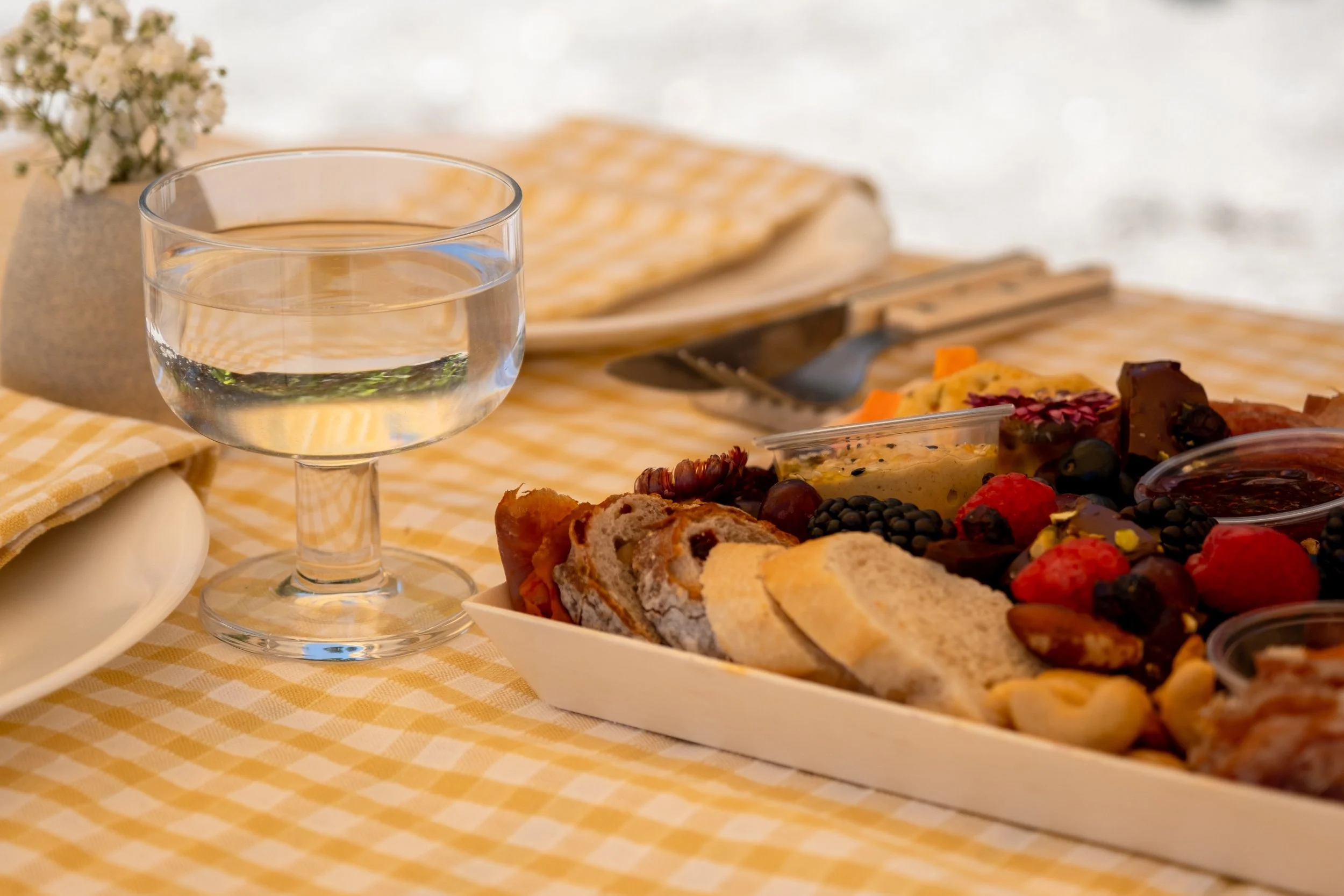 A glass of water, a small flower arrangement, a tray of assorted bread and baked goods, and a variety of berries on a checkered yellow tablecloth set for a meal.