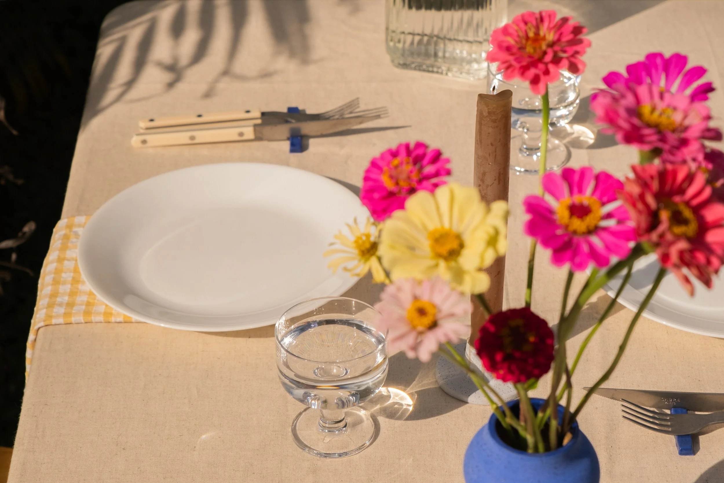A dining table set with a white plate, a glass of water, a flower vase with colorful pink, yellow, and red flowers, a tall candle, cutlery, and a glass jar, with sunlight casting shadows.