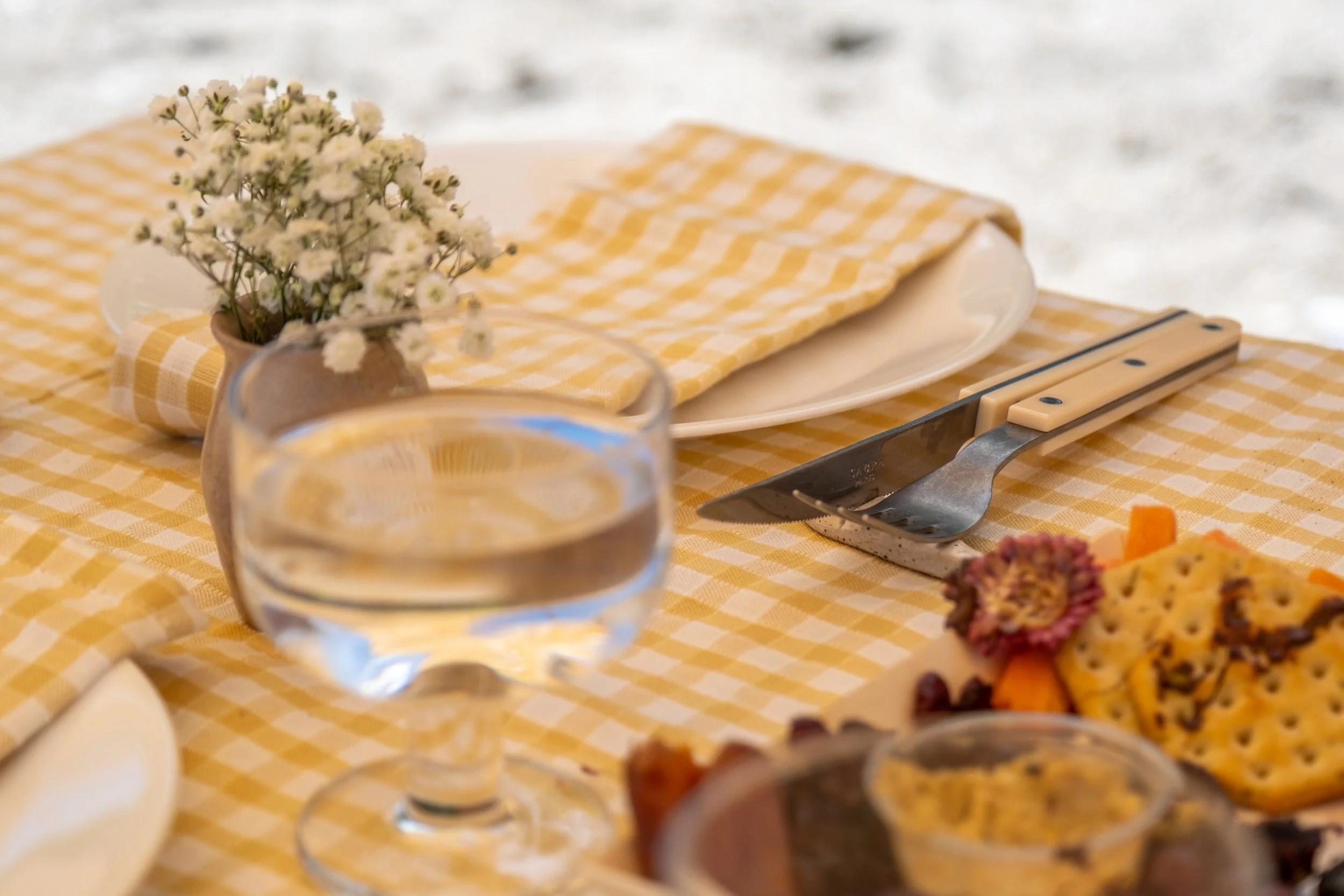 A table set with a glass of water, white plates, a knife and fork, a small vase with white flowers, and a platter of assorted snacks on a yellow and white checkered tablecloth.