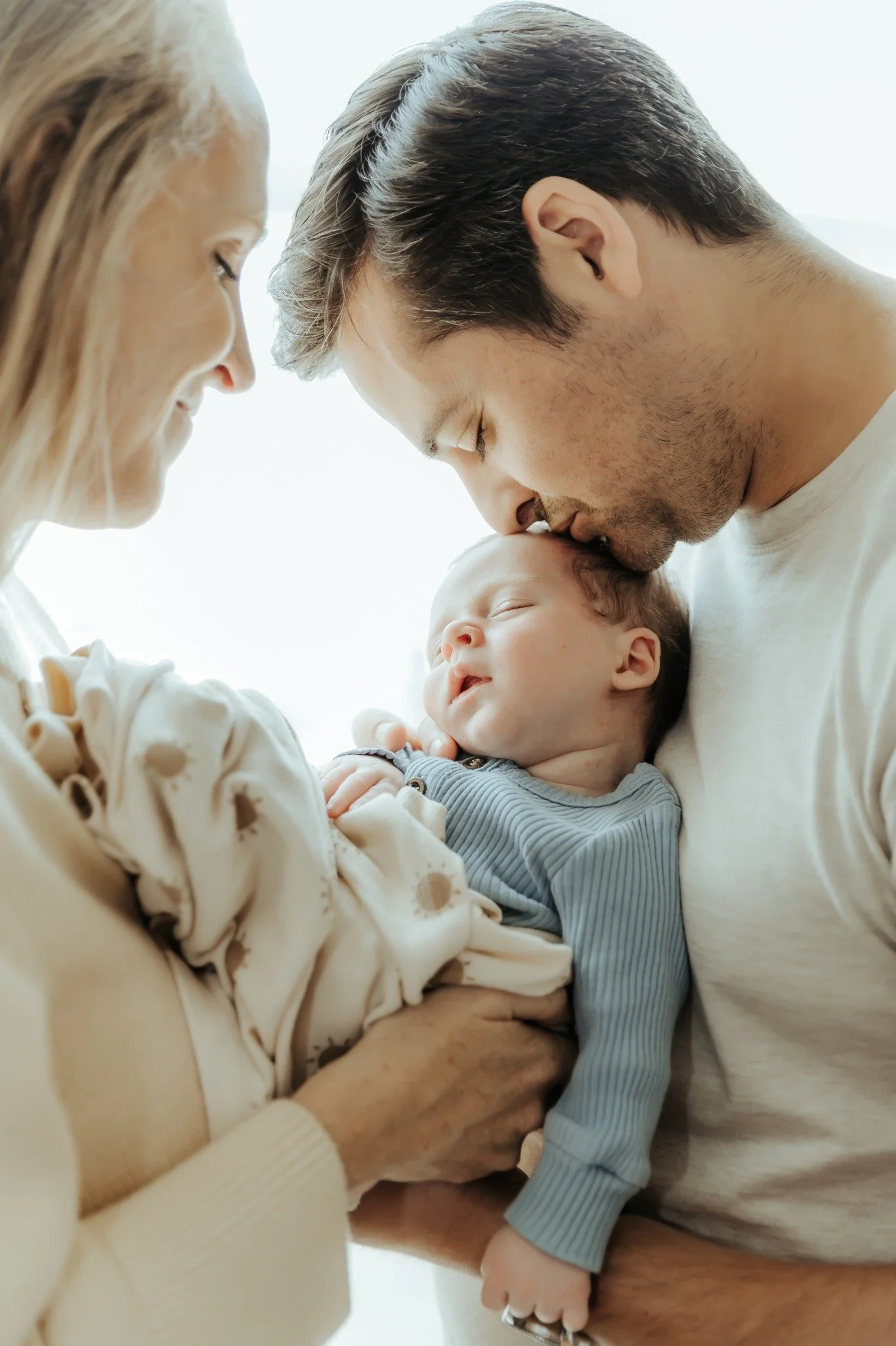 A family holding a sleeping baby, with the father kissing the baby's forehead.