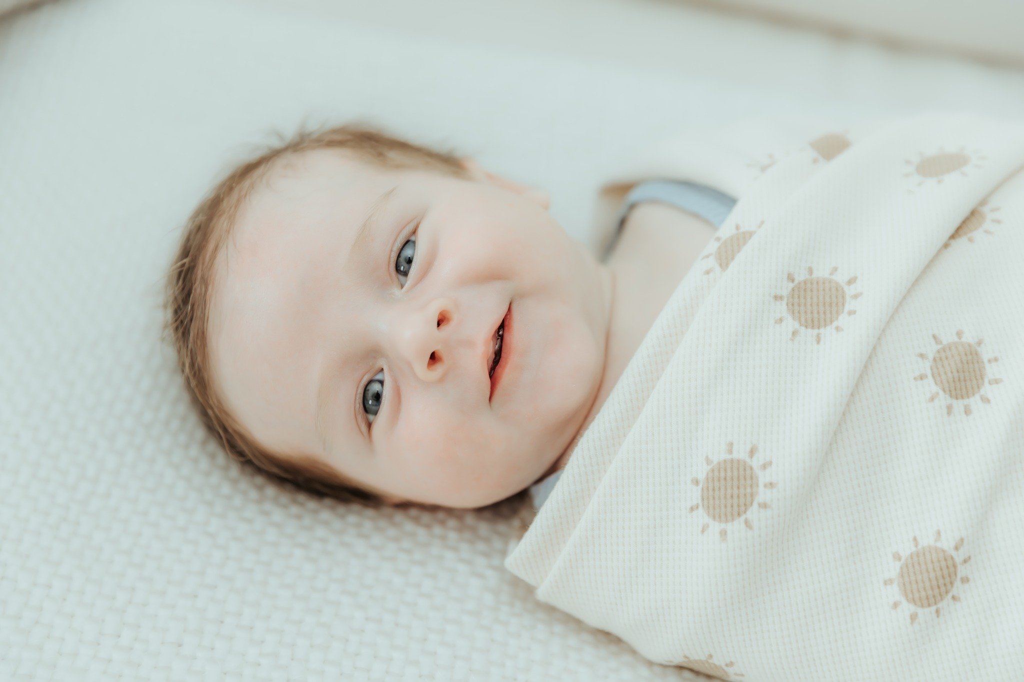 Close-up of a smiling baby lying on a soft surface, wrapped in a cream-colored blanket with sun illustrations.