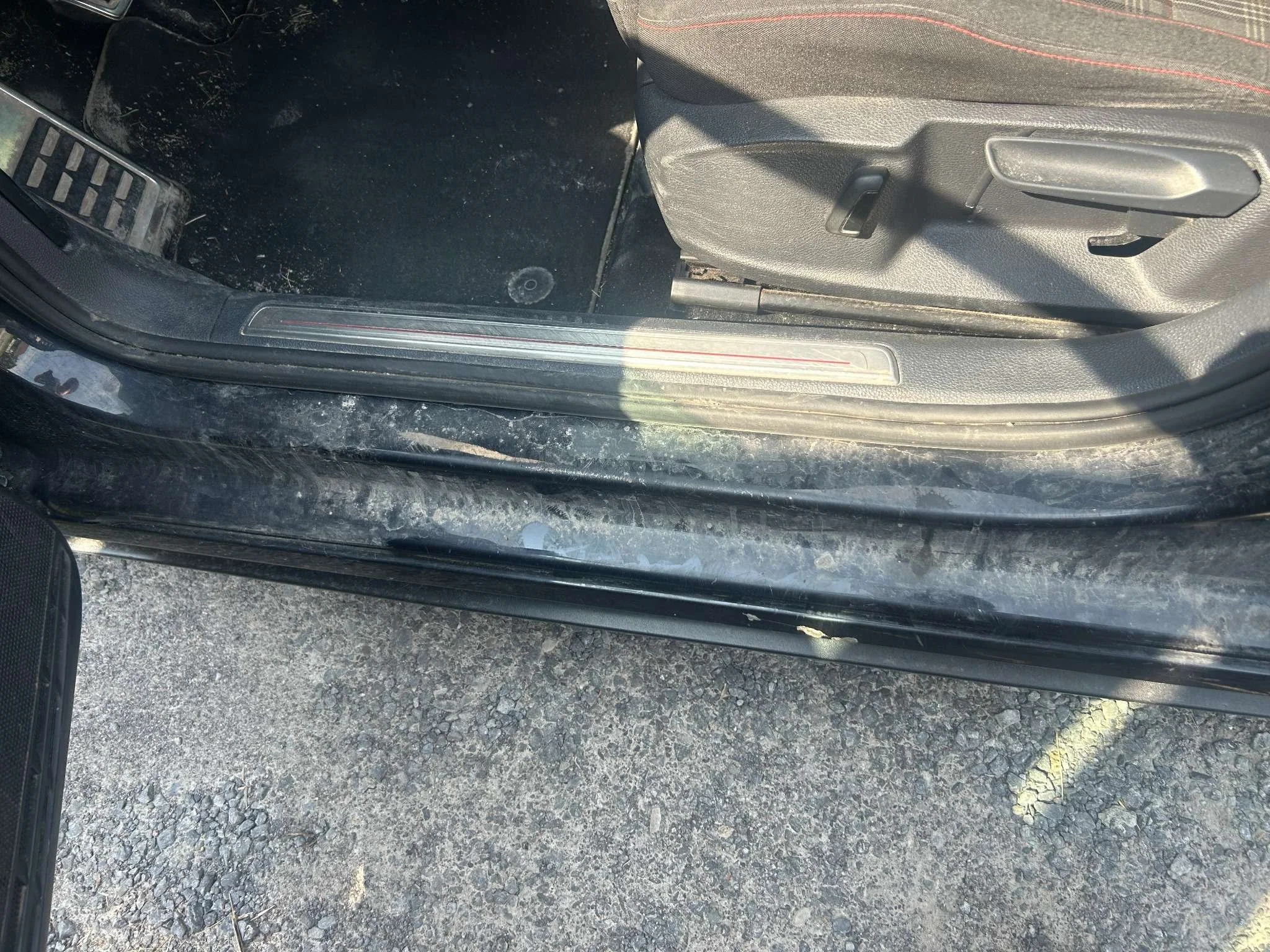 Inside of a black car showing the door frame, floor with dirt, and a partially visible gray seat with red stitching.