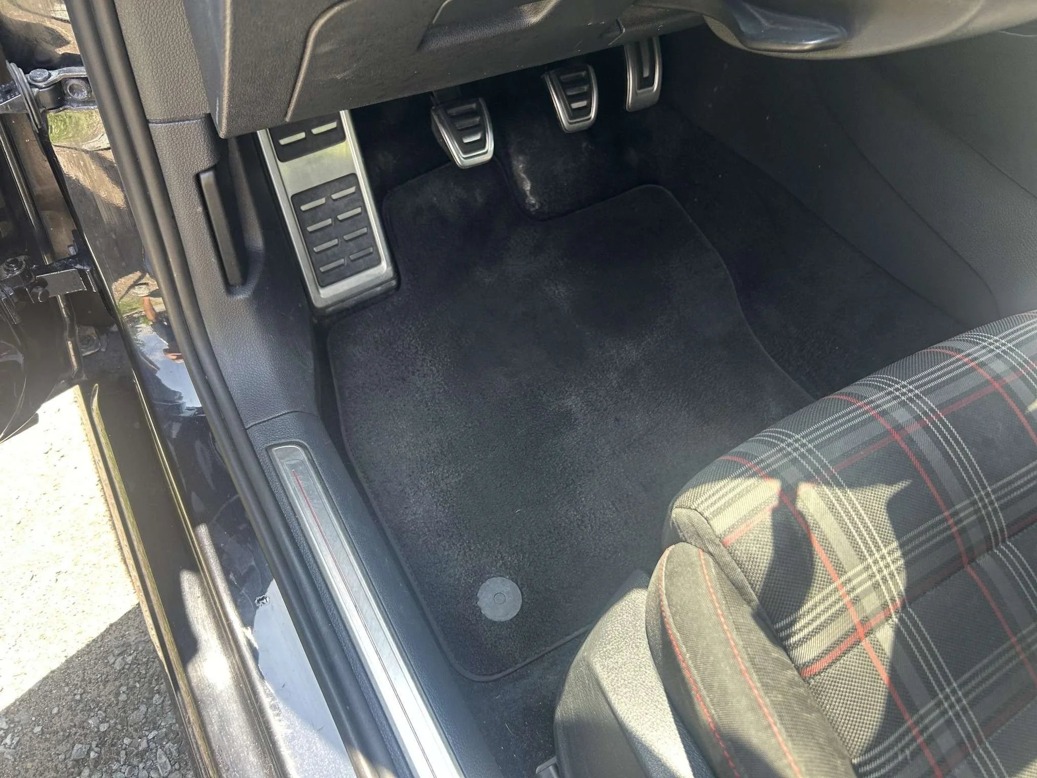 Close-up of the driver's side footwell of a car, showing three metallic pedals and a black carpeted floor mat with a plaid-patterned seat nearby.