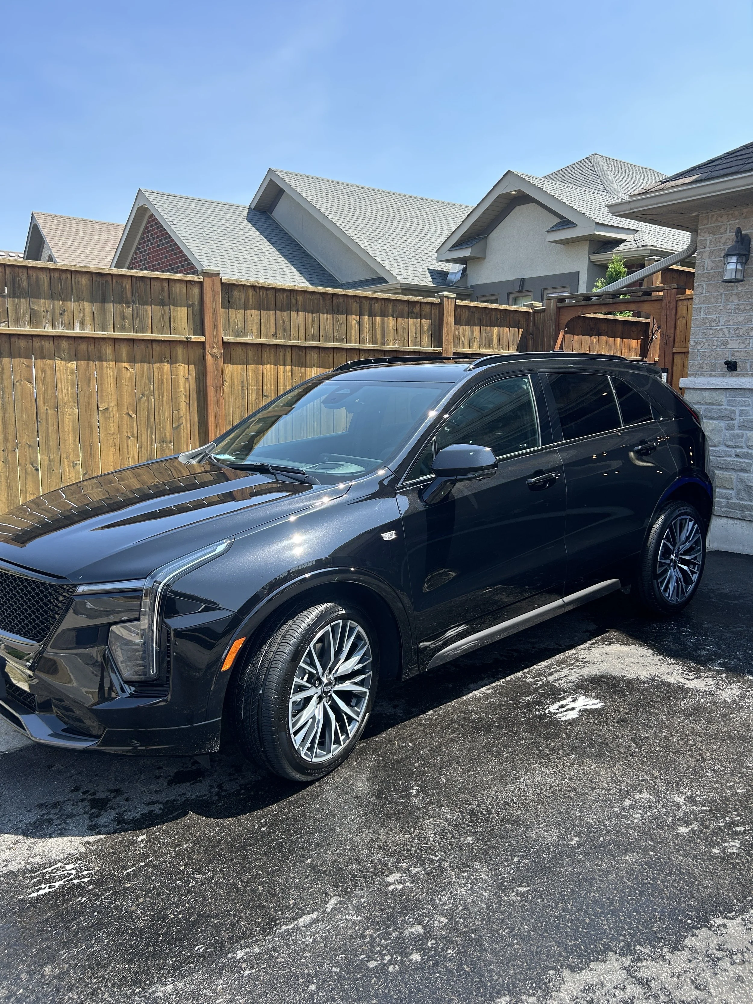 Black luxury SUV parked on a driveway in front of a wooden fence, with a suburban house and blue sky in the background.