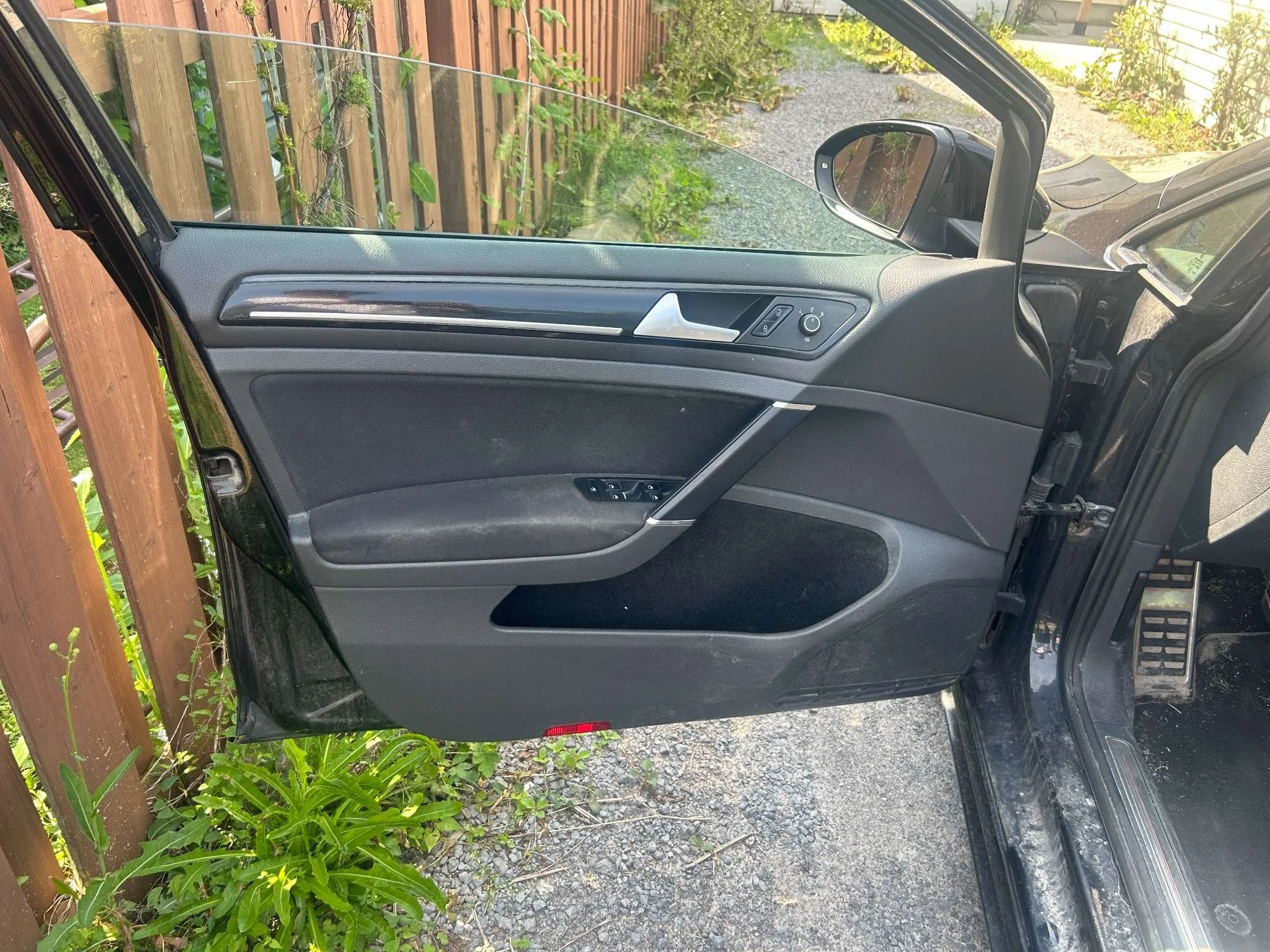 The image shows the interior side of a car door that is open, revealing the window, door handle, and control buttons, with a gravel driveway and some greenery visible outside.