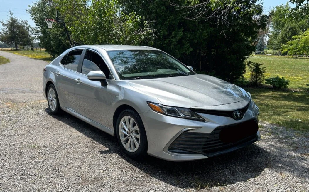 Silver Toyota sedan parked on a gravel driveway with green trees and a basketball hoop in the background.