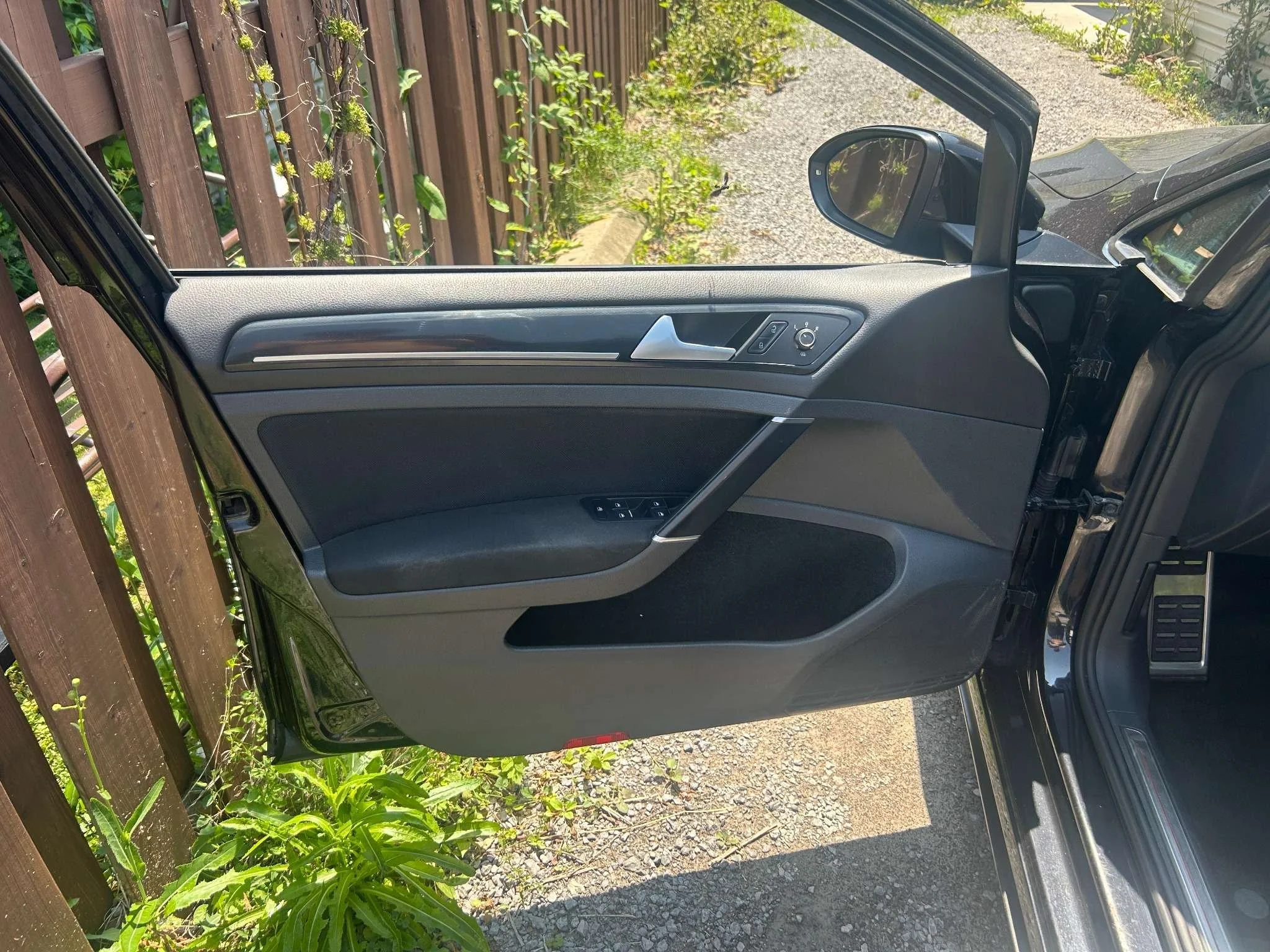 Open car door inside view of the driver side with window controls and side mirror, parked next to a wooden fence and greenery.