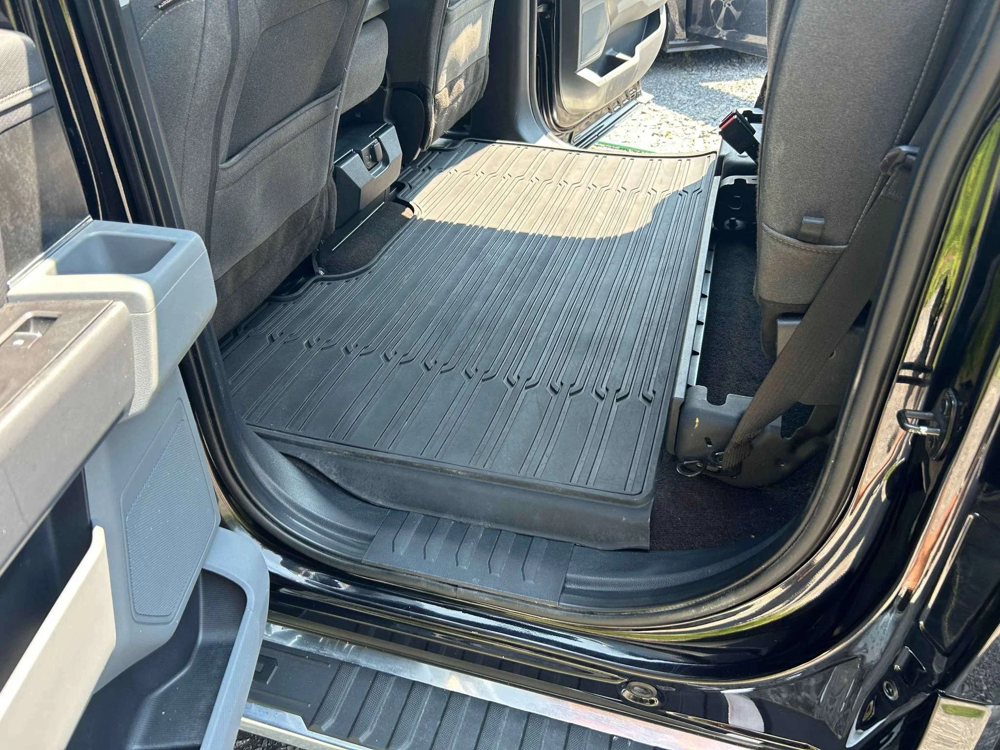 Interior of a vehicle showing a black rubber cargo liner on the floor behind the front seats, with part of the open vehicle door visible.