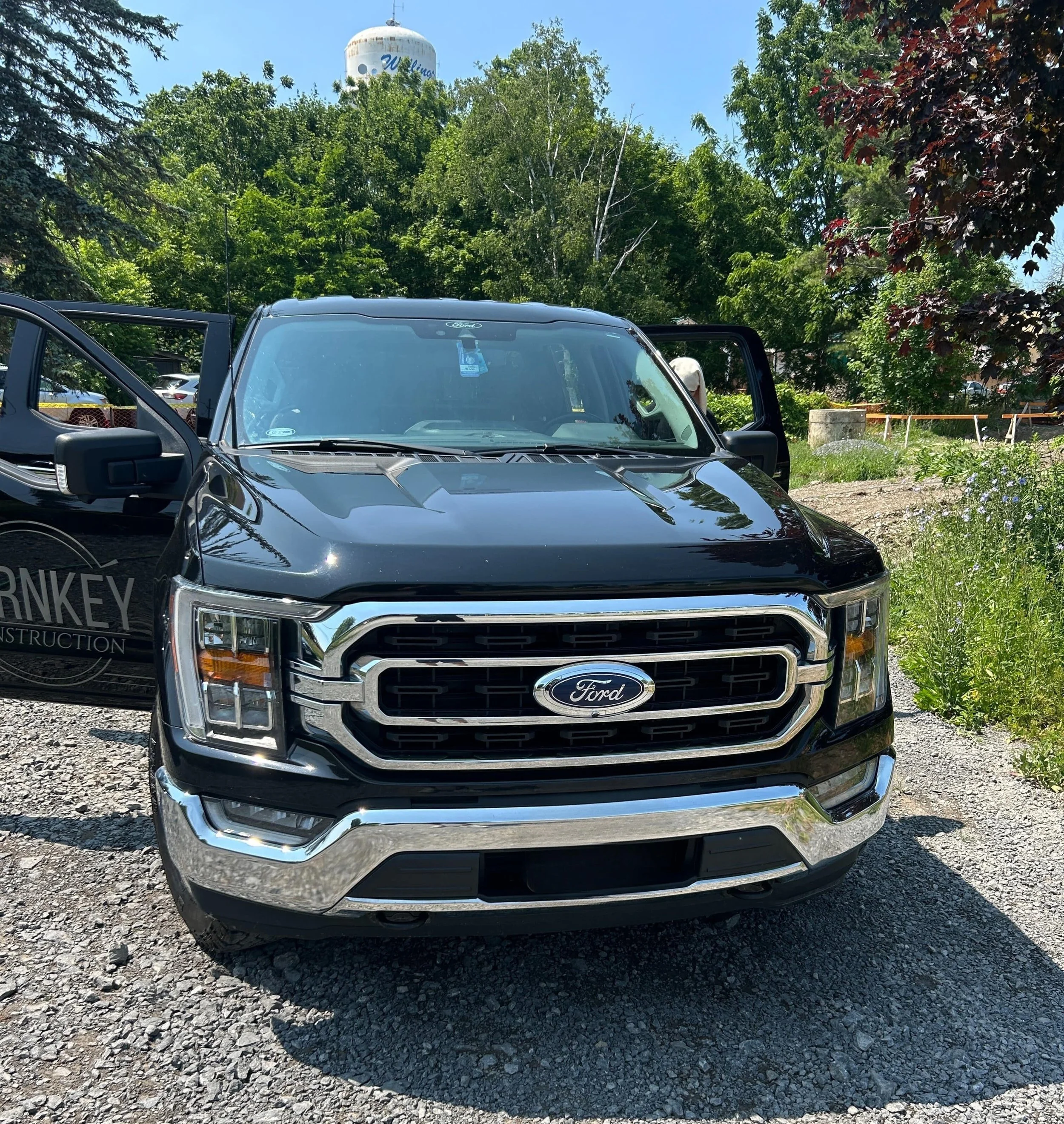 A black Ford pickup truck parked outdoors on a gravel surface with green trees and a water tower in the background.