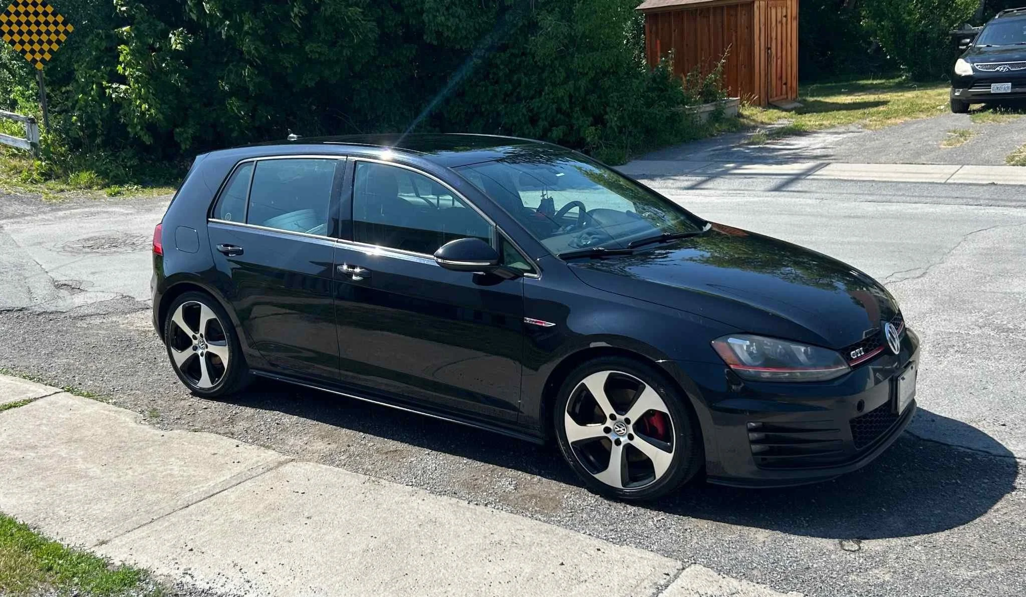 Black Volkswagen GTI hatchback parked on a street near a sidewalk, with trees and a wooden fence in the background.
