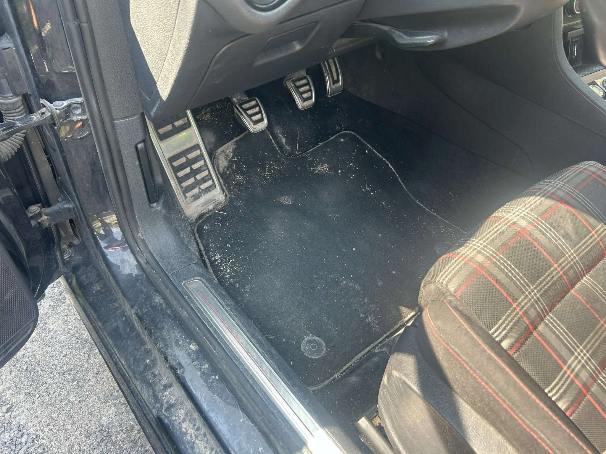 The driver side floor of a car showing dirt and debris on the black carpet, the pedals, and part of the driver's seat with plaid upholstery.