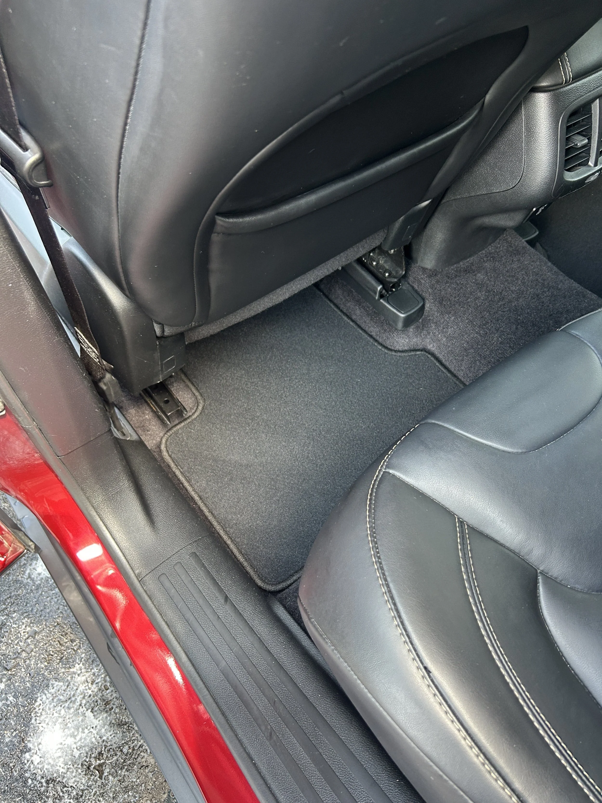 The back seat floor area of a vehicle with black carpeting, a black leather seat, and the back of the front seat with a storage pocket.