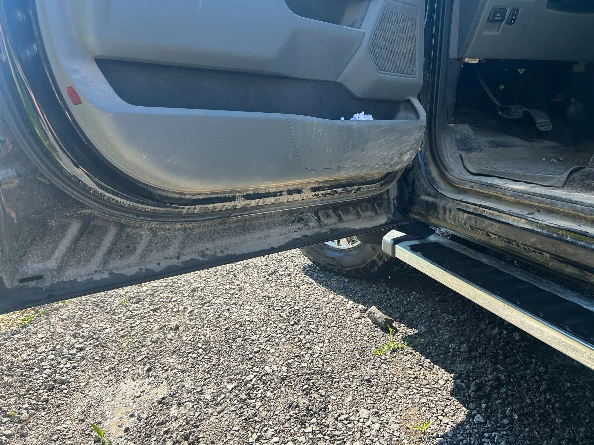Close-up of the interior of a vehicle's driver's side door and footwell area, showing dirt and a running board step outside.