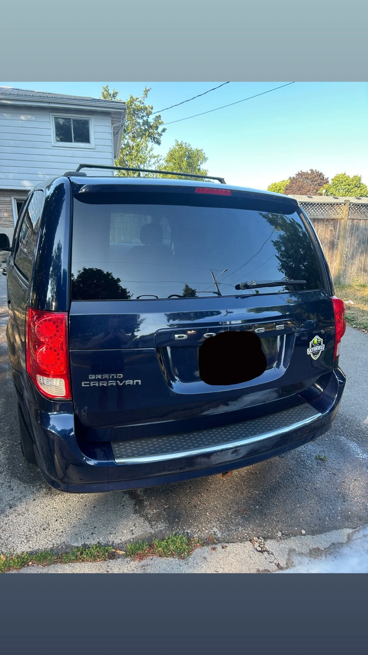 Blue Dodge Grand Caravan minivan parked in a driveway, with a house in the background and a Skyhawks sticker on the rear window.