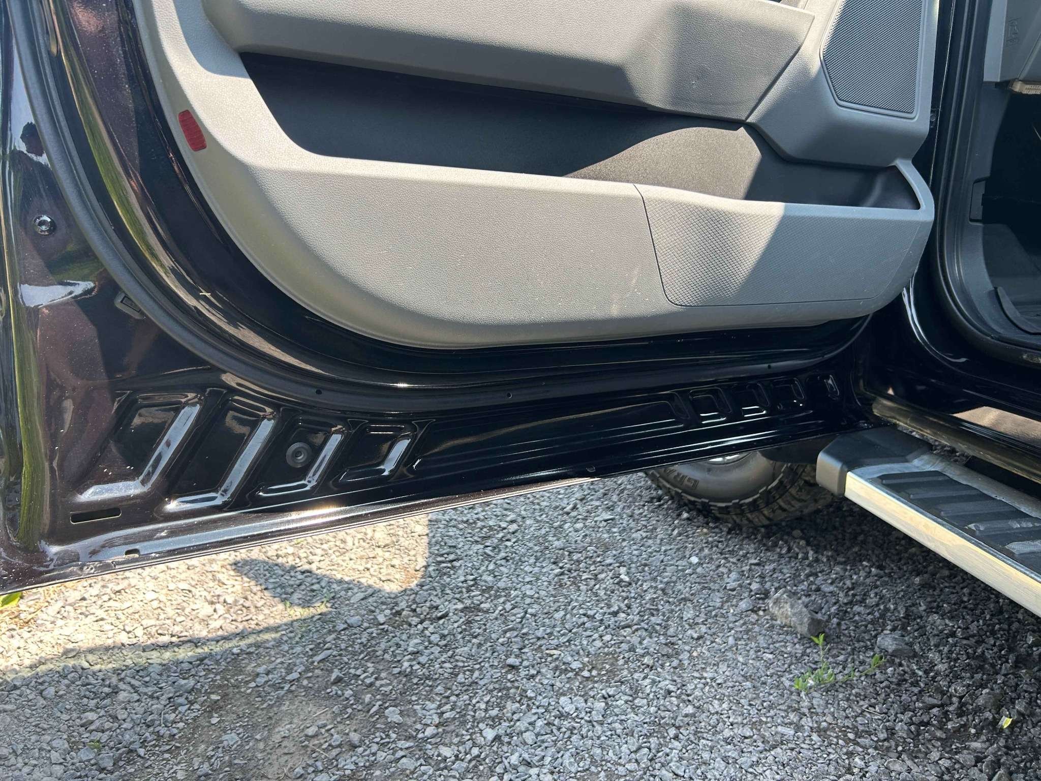 Close-up of the interior of a vehicle's door, showing the lower part with a black metal frame, gray plastic panel, and a textured step area, with a gravel ground outside.