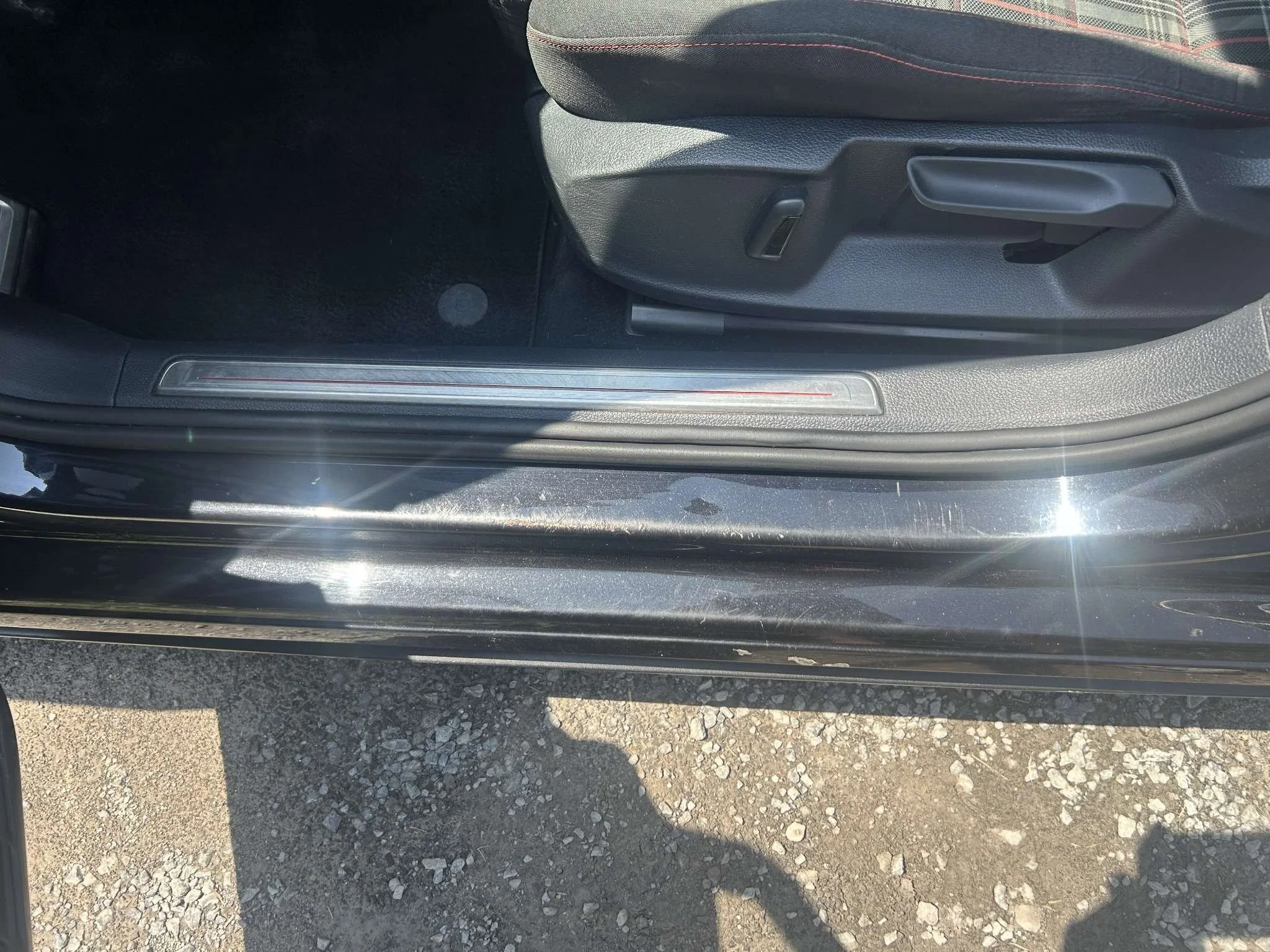 Close-up of the lower part of a car door frame and a partially open car door, showing the interior floor mat and seat adjustment controls. The car's exterior is reflecting sunlight.