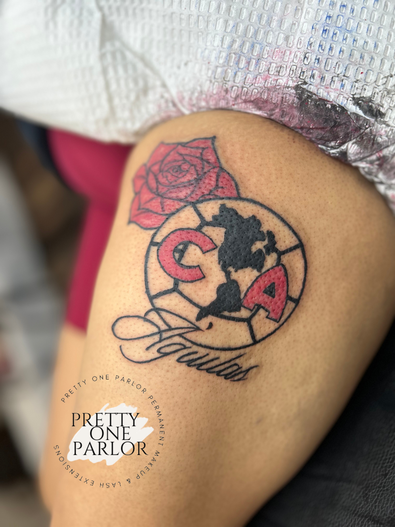 Tattoo on a person's upper arm featuring a red rose, a circular design with black map outlines, the letters C and A in red, the word "Fabulous" in cursive, and a Pretty One Parlor logo with text around a white paint splash.