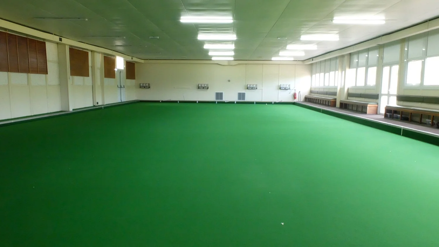 Indoor sports room with green artificial turf, benches along the side, and large windows. A lawn bowling green.