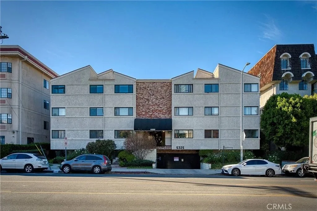 Multi-story residential building with a brick and stucco facade, parked cars in front, and a clear blue sky.