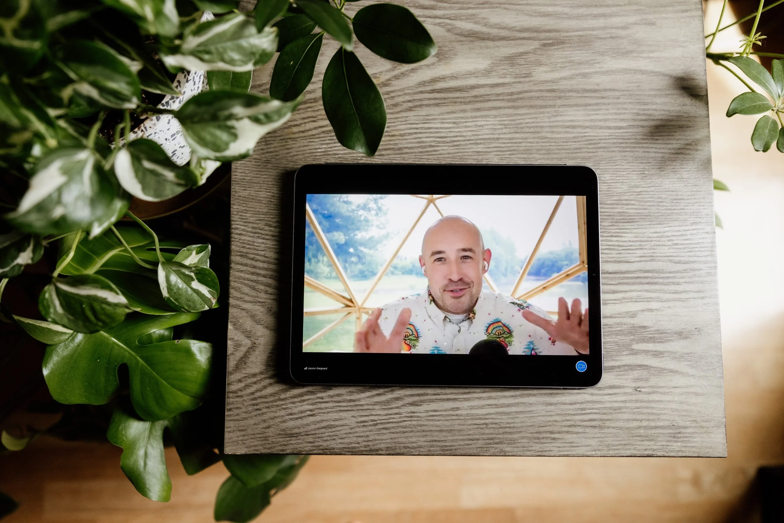 A tablet displaying a man with a bald head and white shirt, talking with hand gestures, with green plants on a wooden table.