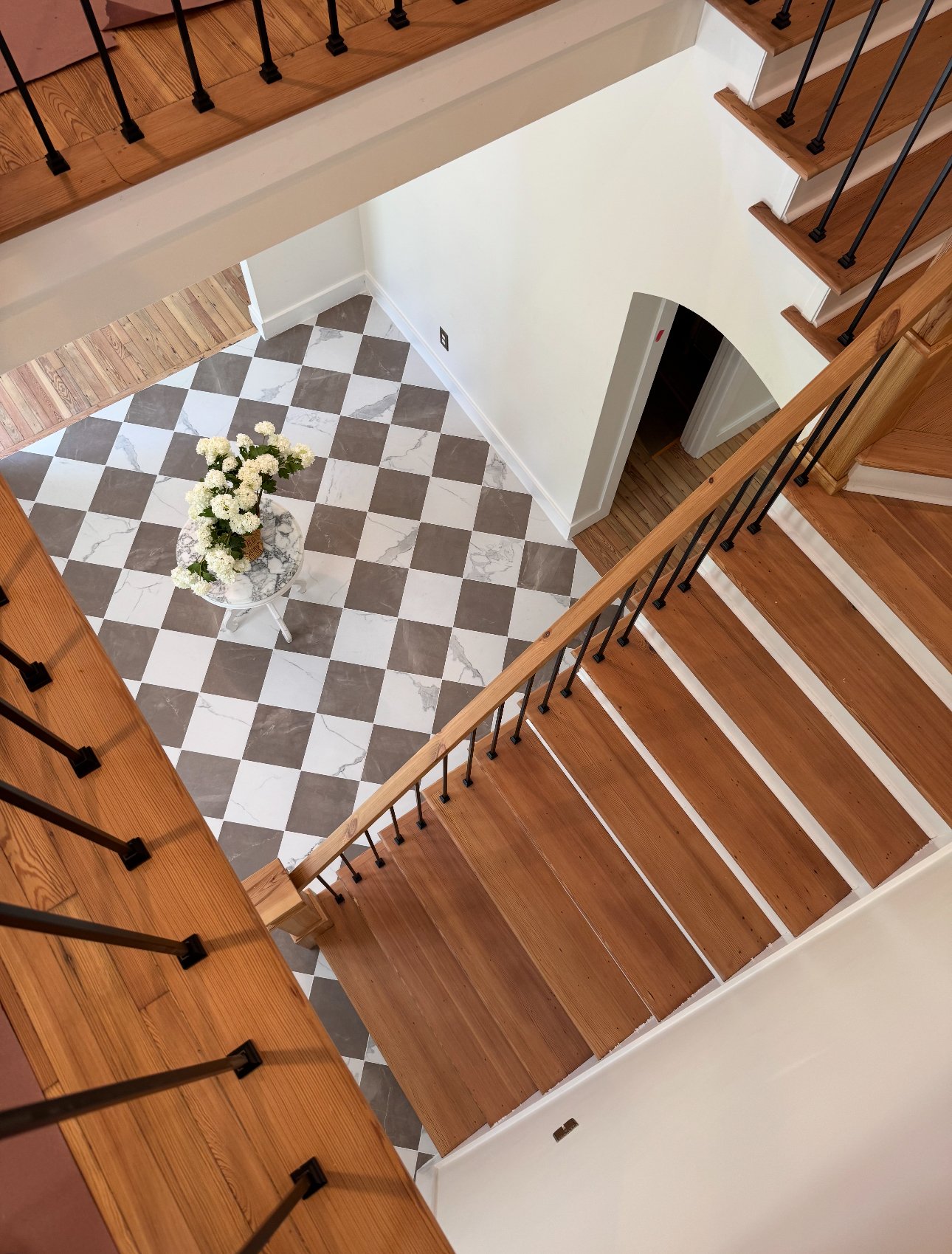 An overhead view of a wooden staircase with black metal railings leading to a tiled entryway with a small round table holding a vase of white flowers.