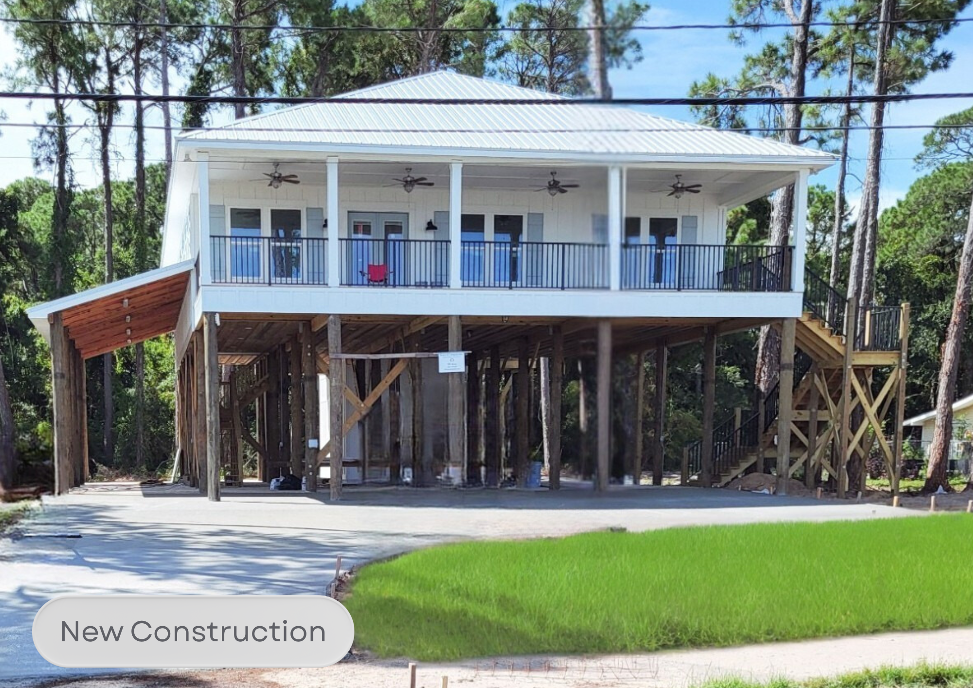 A new elevated house under construction with a white exterior, metal roof, and a large wrap-around porch with ceiling fans. The foundation is incomplete with visible support beams and wooden framework, and stairs lead up to the porch. There is a gras
