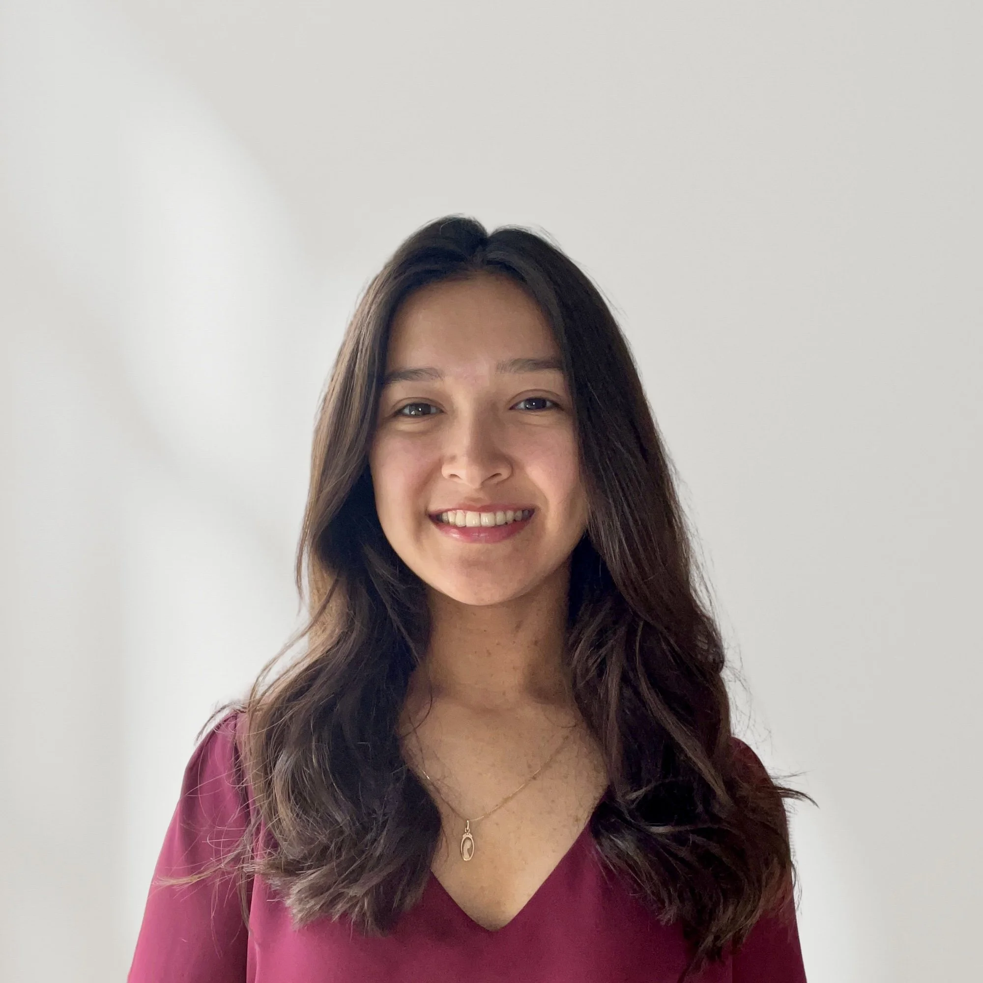 A young woman with long dark hair smiling, wearing a maroon top and a gold necklace, standing against a plain white wall.