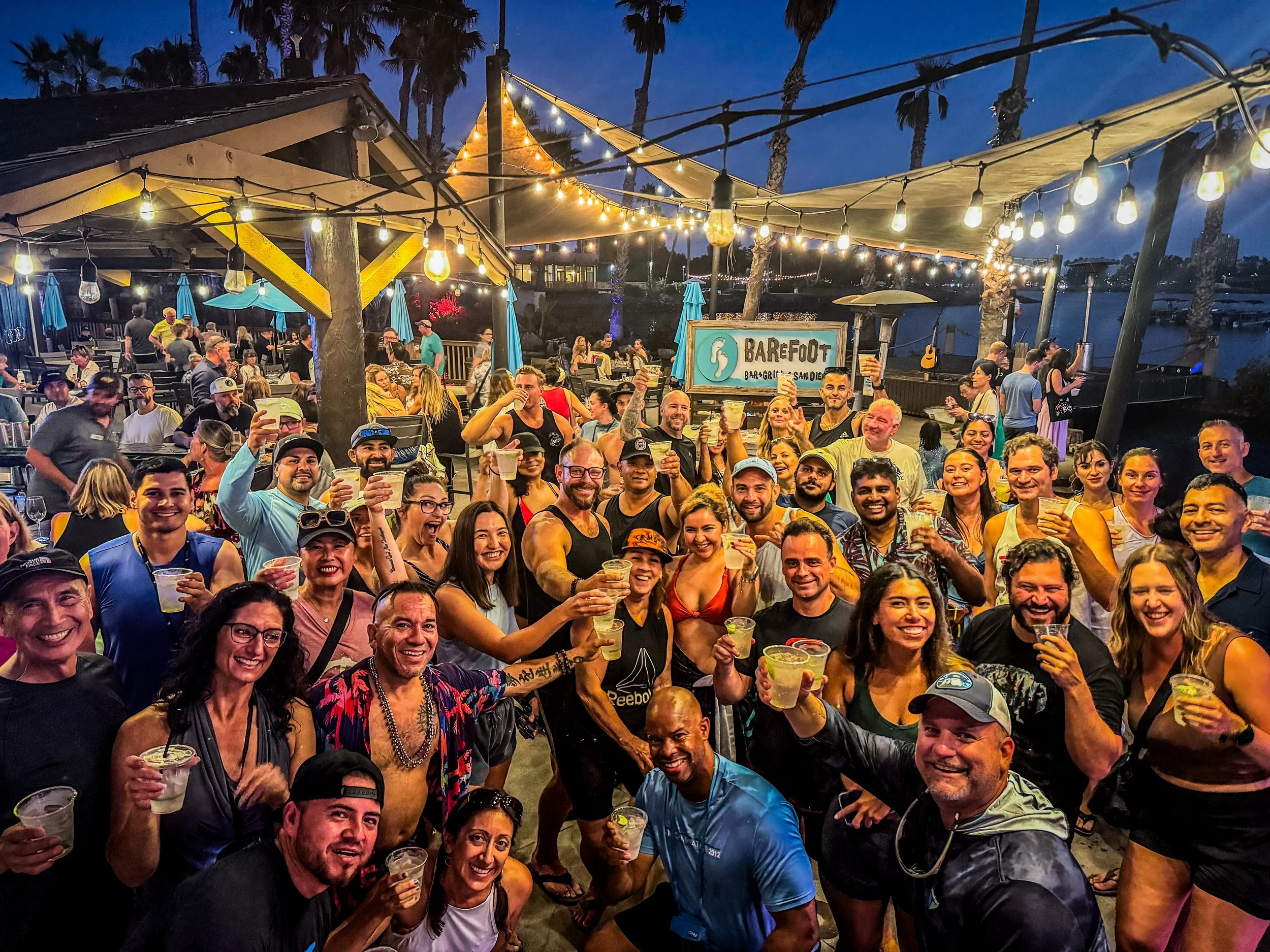 Paddle Boarding SD Paddlers at the Barefoot Bar. A large group of diverse people celebrating outdoors during the evening at a bar or restaurant with string lights and palm trees, holding drinks and smiling.