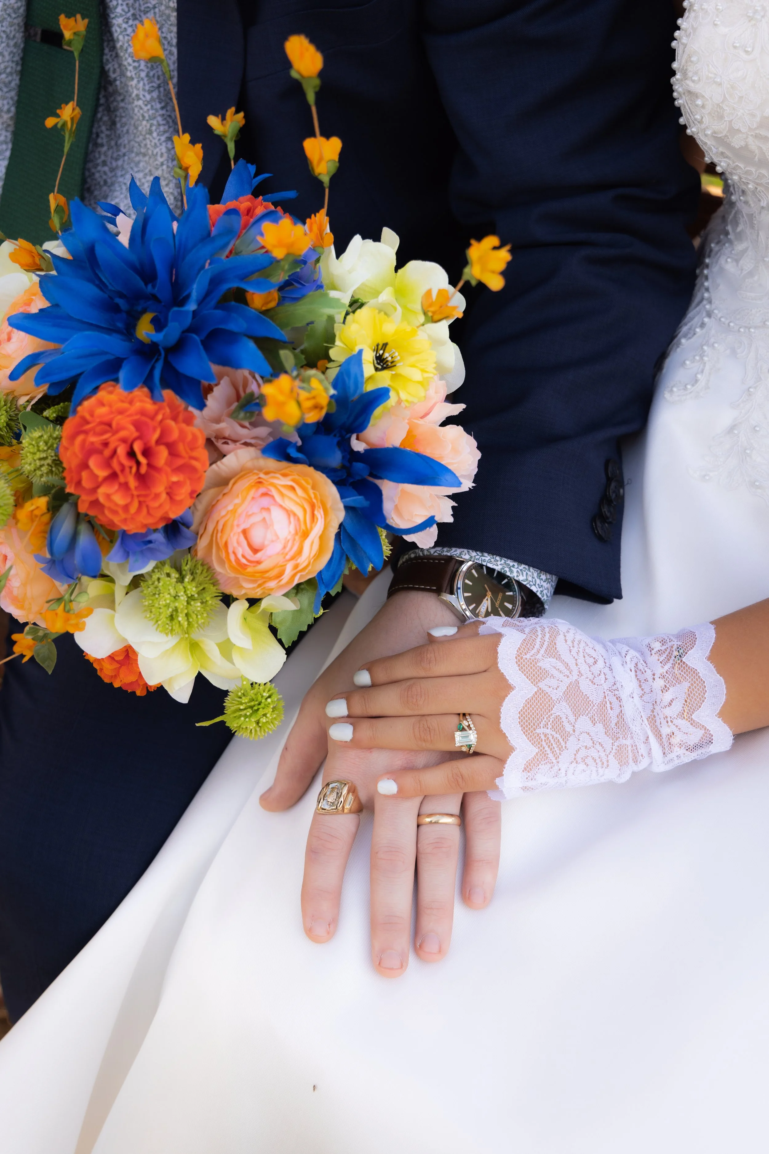 Close-up of a bride and groom's hands showing rings and lace gloves, with the bride's hand resting on the groom's hand, holding a colorful bouquet of flowers, against a backdrop of wedding attire.