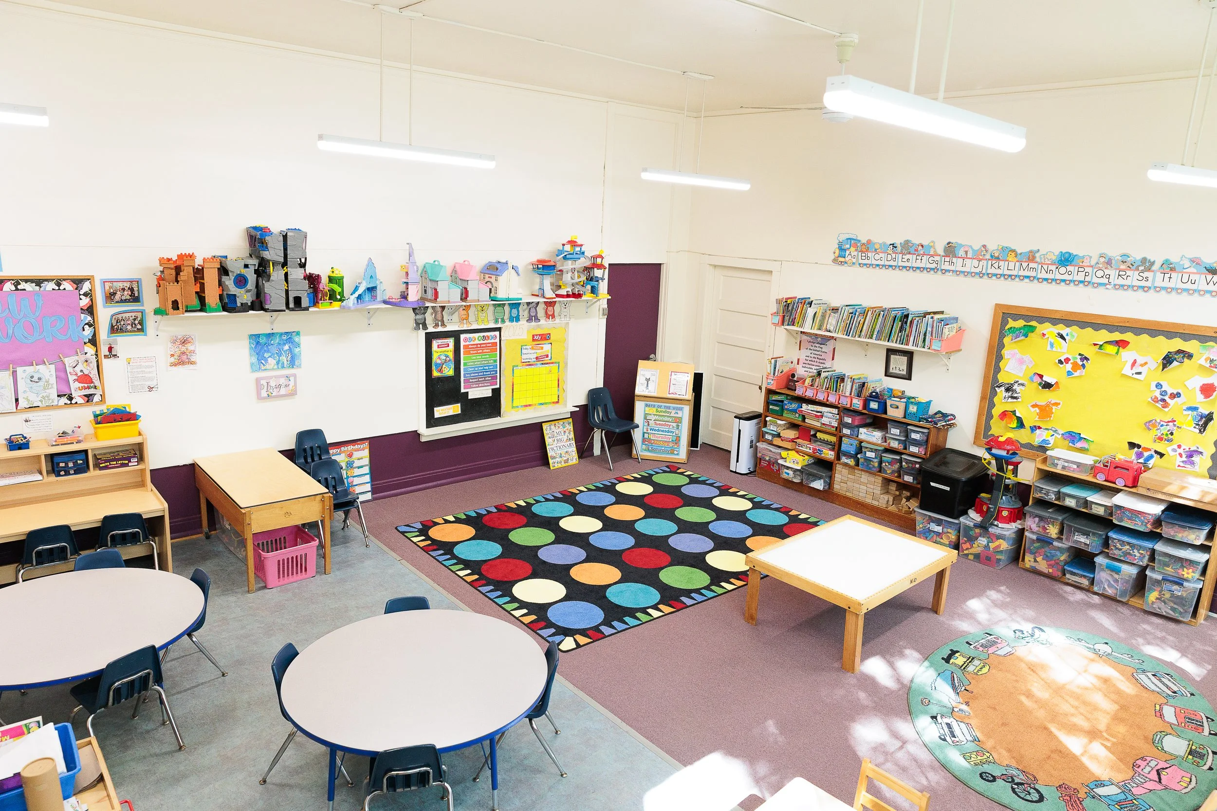 Interior of a colorful preschool classroom with tables, chairs, shelves of books, and educational decorations.