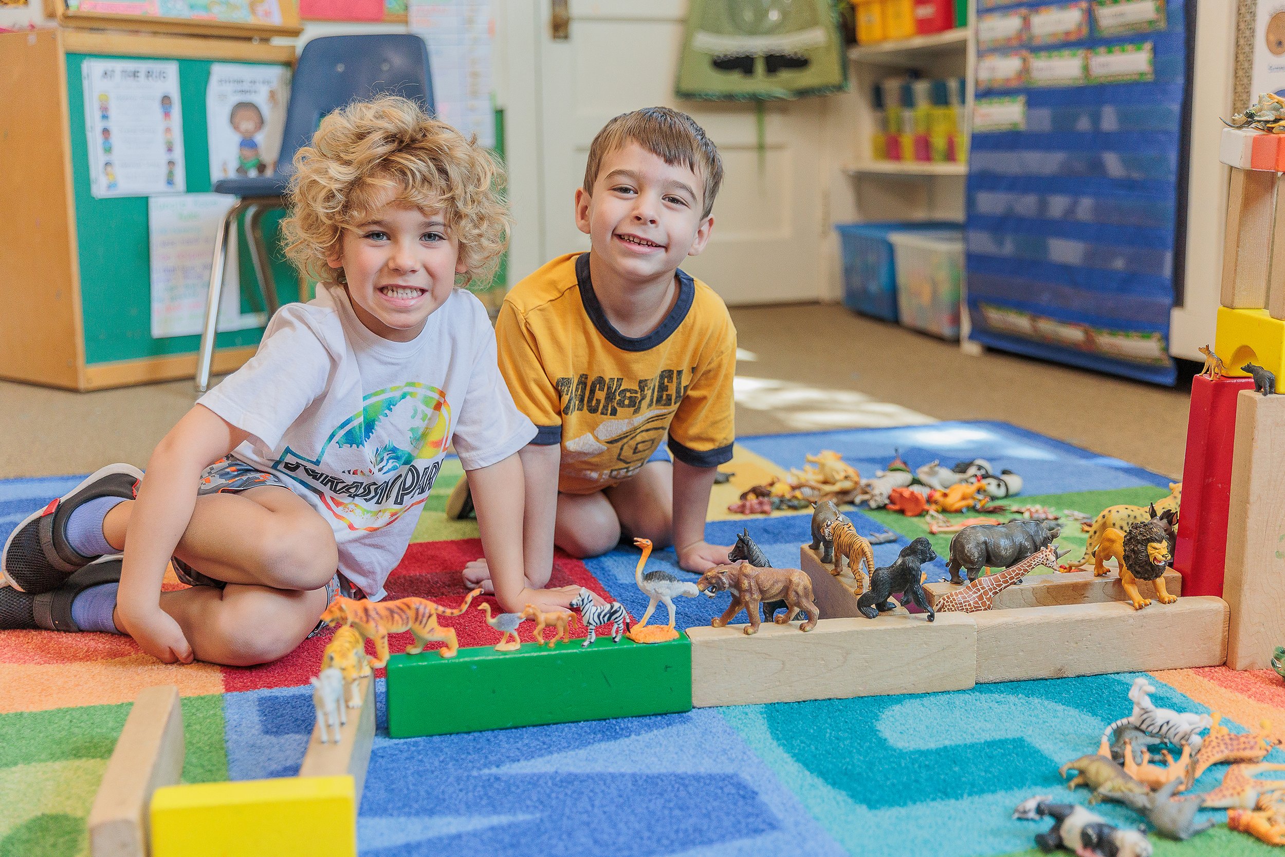 Two young boys playing with toy animals on a colorful carpet in a classroom.