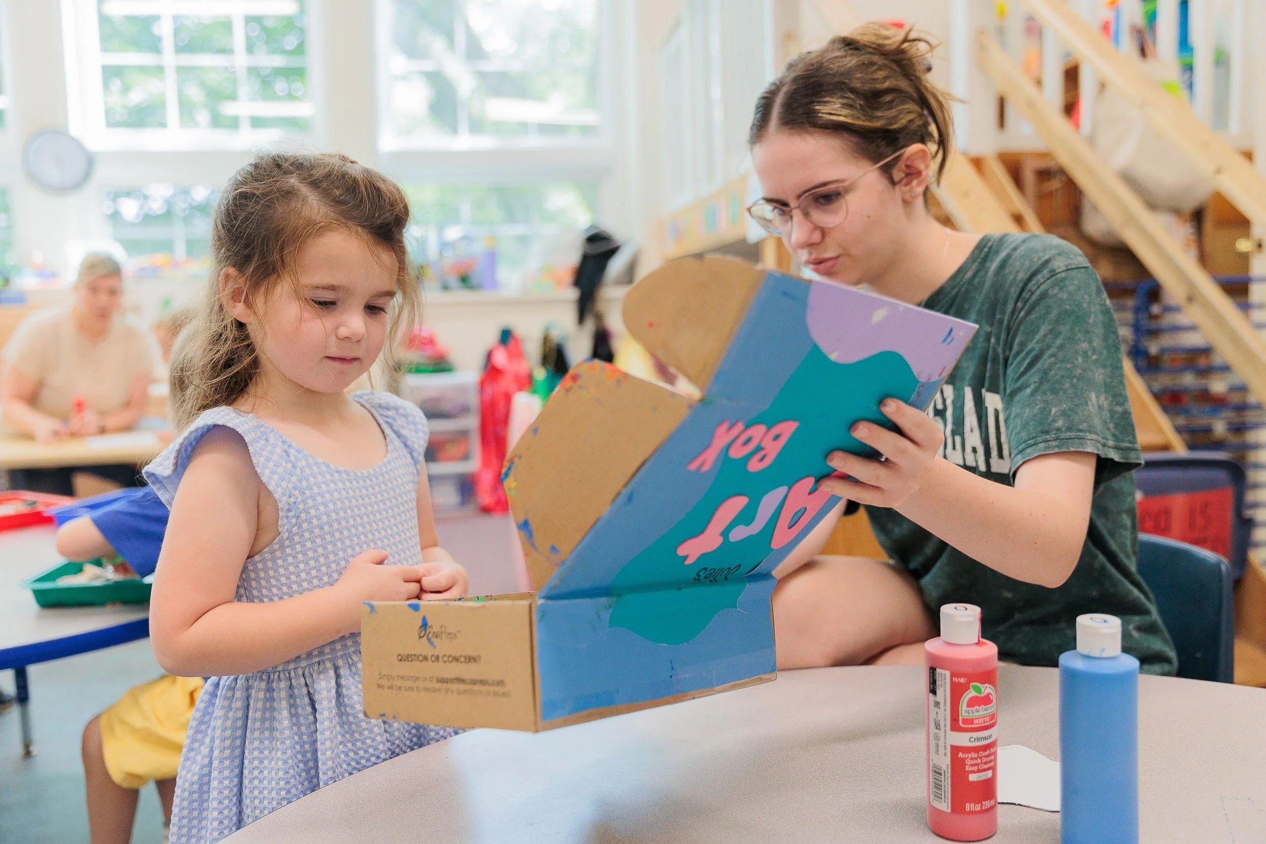 A young girl and an employee are sitting at a table in a classroom, looking at a cardboard box with colorful designs.