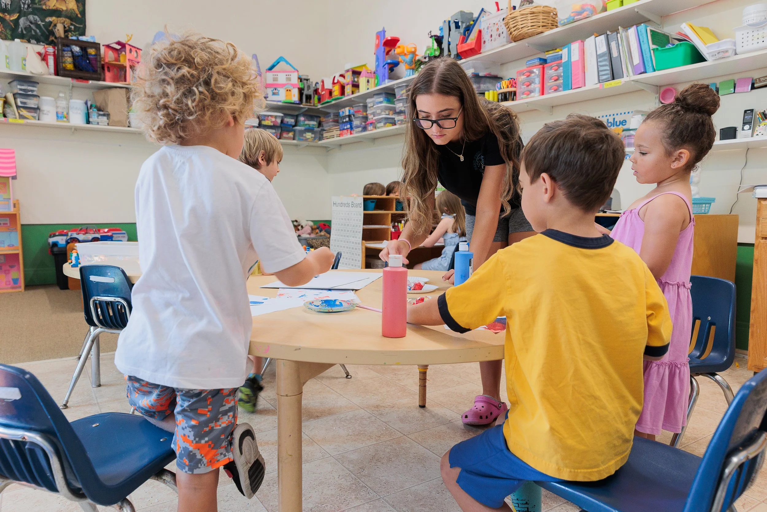 Kids painting at a classroom table with a teacher supervising.