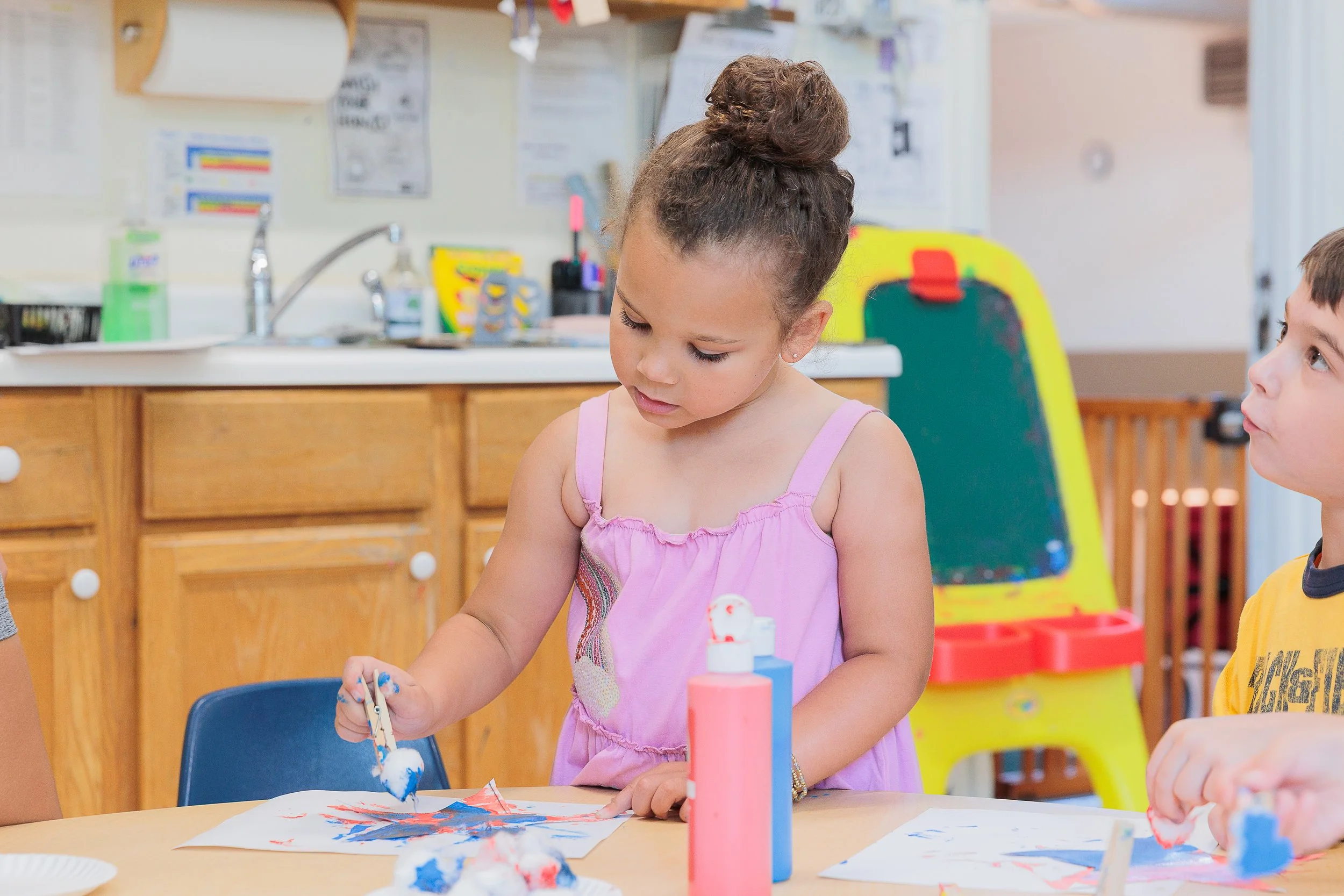 Young girl painting with blue and red paint on paper at a table, with children nearby in a classroom setting.