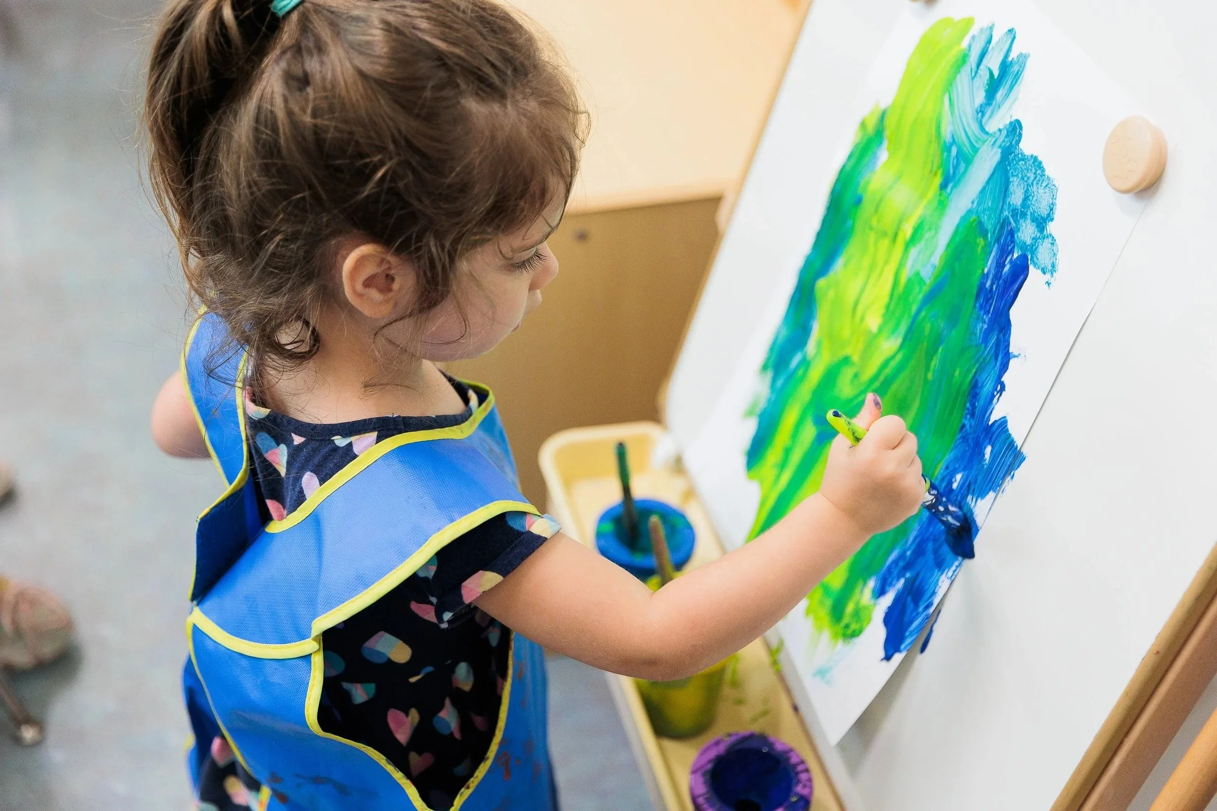A young girl with curly brown hair, wearing a blue apron, is painting with green and blue colors on a large white canvas attached to an easel.