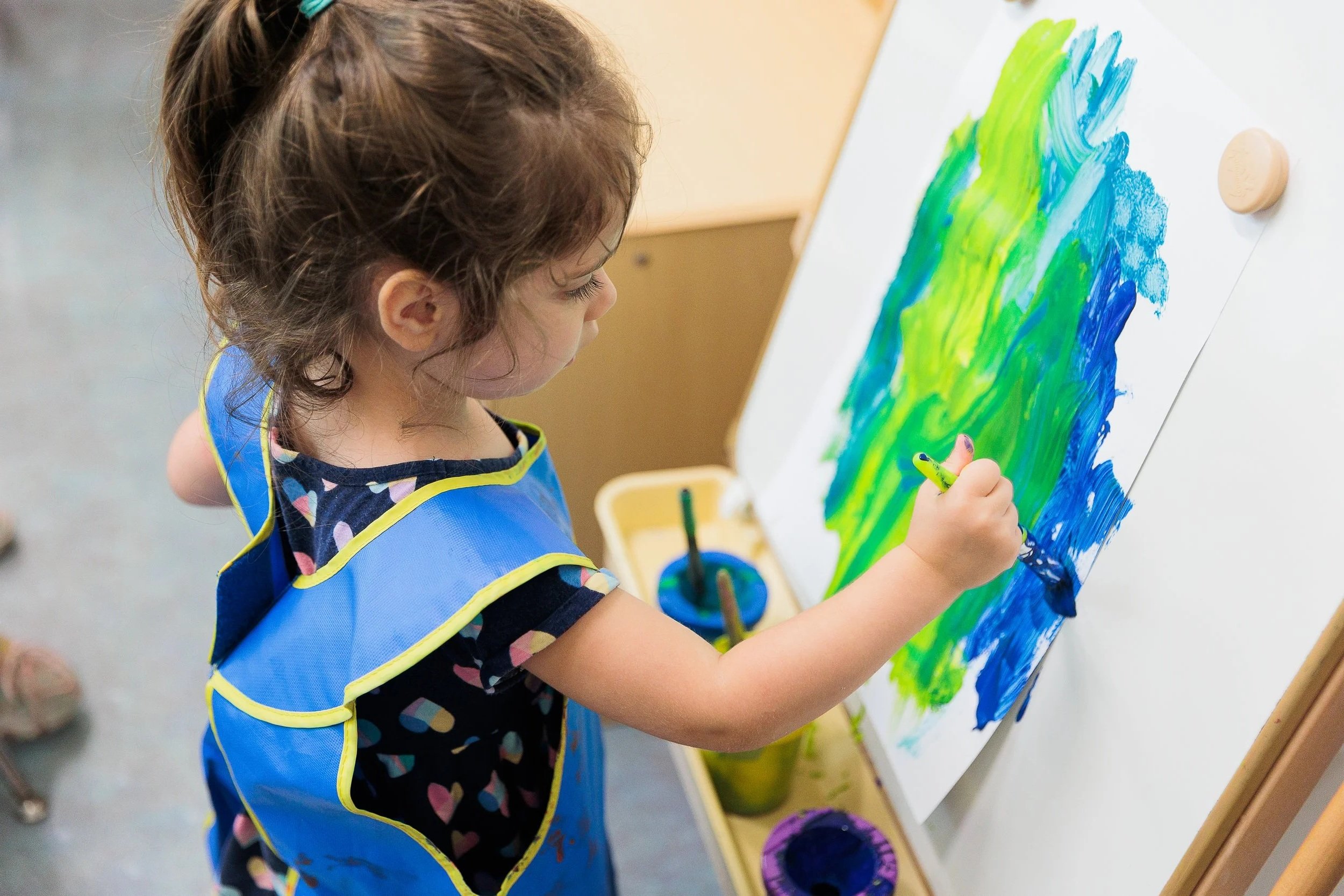 A young girl with curly brown hair, wearing a blue apron, is painting with green and blue colors on a large white canvas attached to an easel.