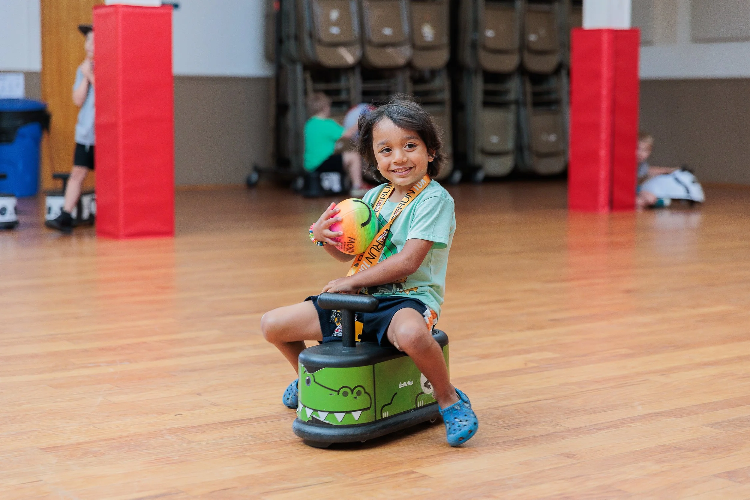 A young boy sitting on a green ride in an indoor gym, holding a colorful ball and smiling at the camera.