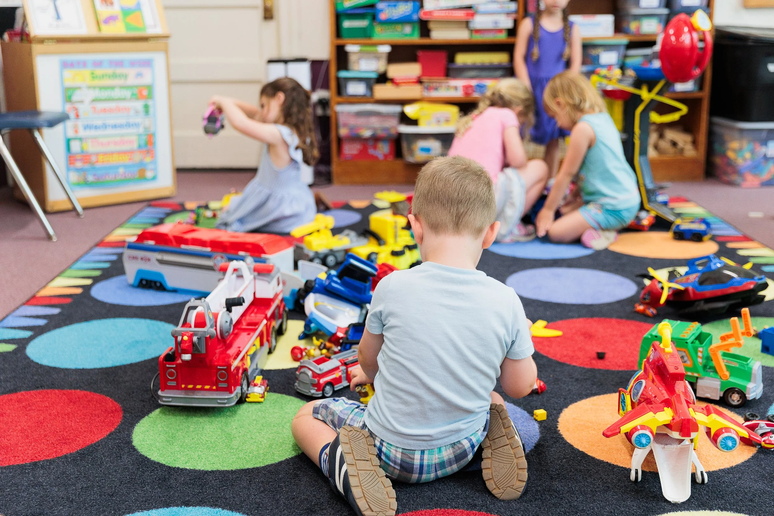 Children playing with toy cars and trucks on a colorful rug in a classroom.