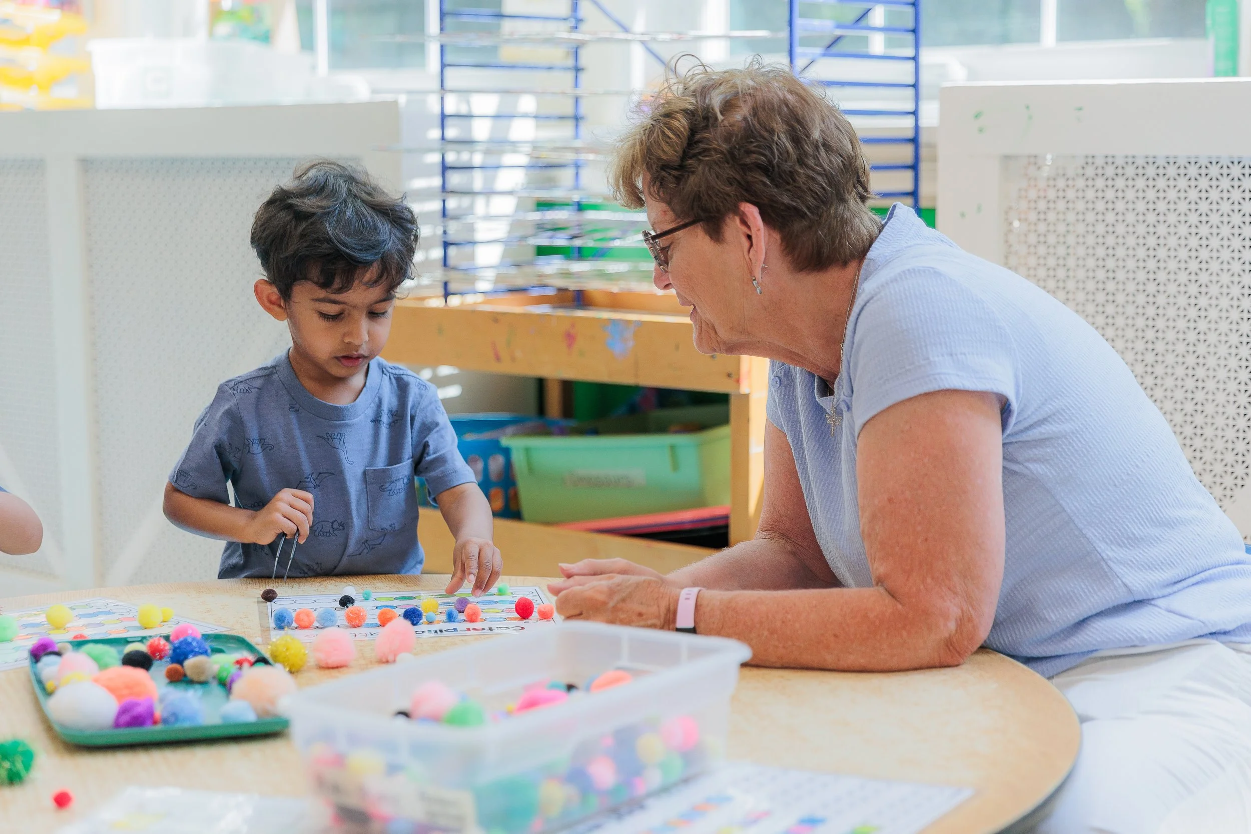 A young boy and an older woman are engaging in a craft activity at a table filled with colorful pom-poms and craft supplies. They are working closely together indoors, in a bright, child-friendly environment.