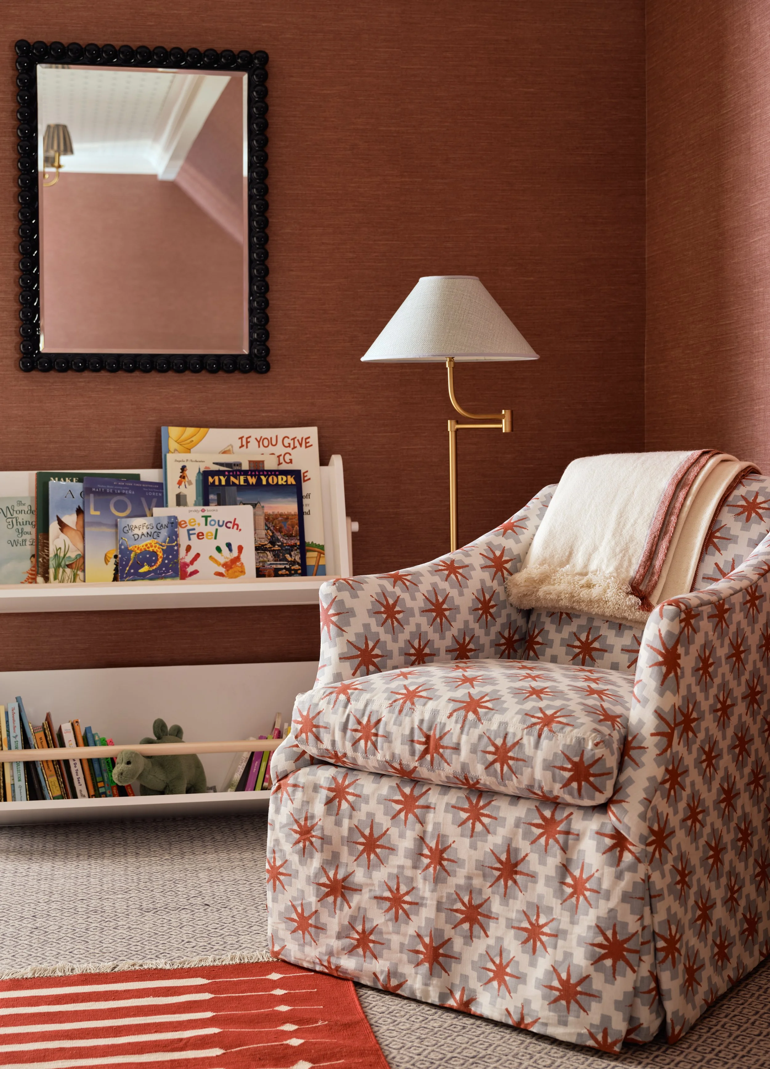 A cozy reading nook featuring an armchair with a star pattern, a cream-colored towel draped over it, a floor lamp, a bookshelf with children's books and a stuffed animal, a mirror on the wall, and a red-patterned rug.
