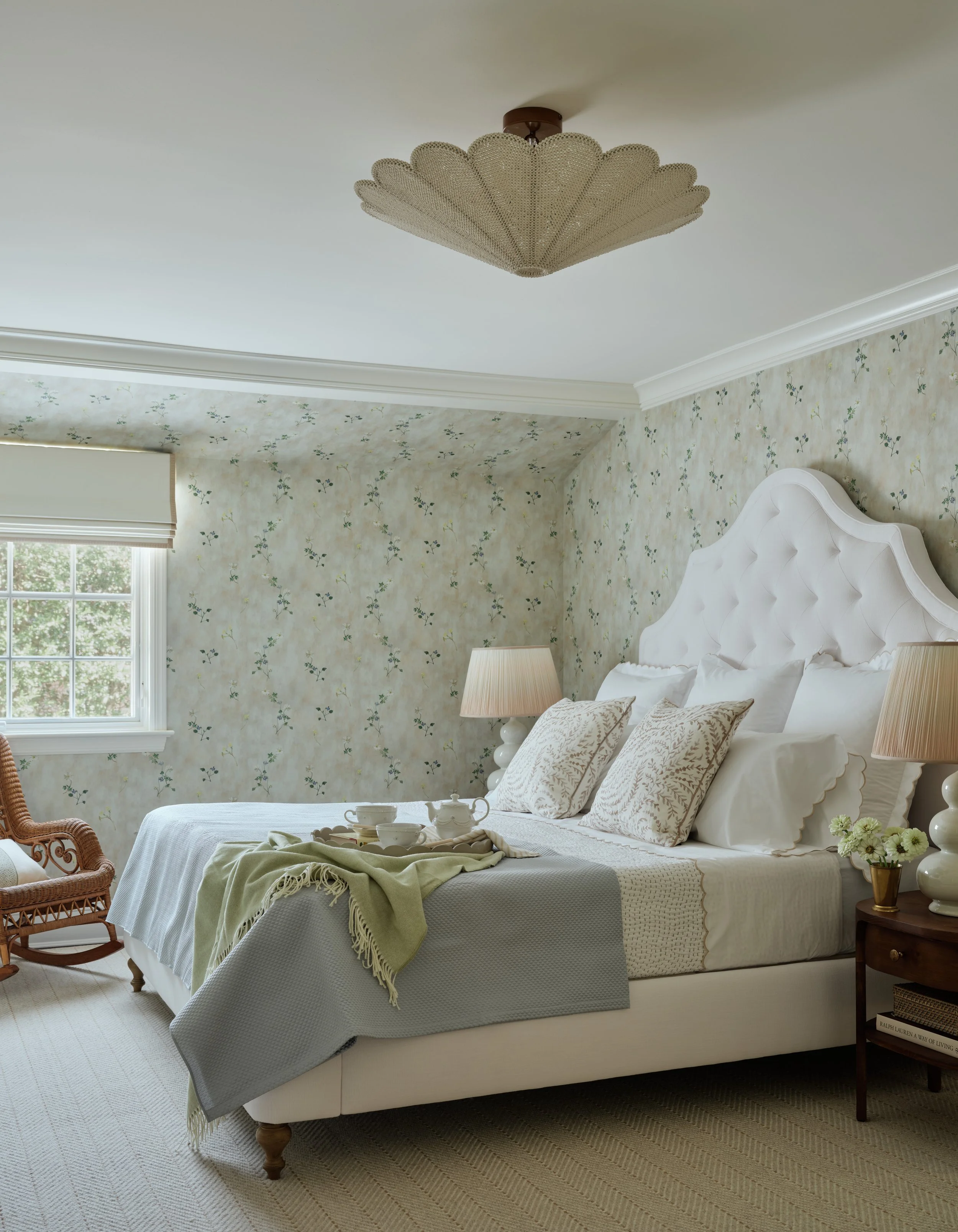 A cozy bedroom with a white bed, floral wallpaper, two lamps, a wicker chair, a window, and a tray with tea set on the bed.
