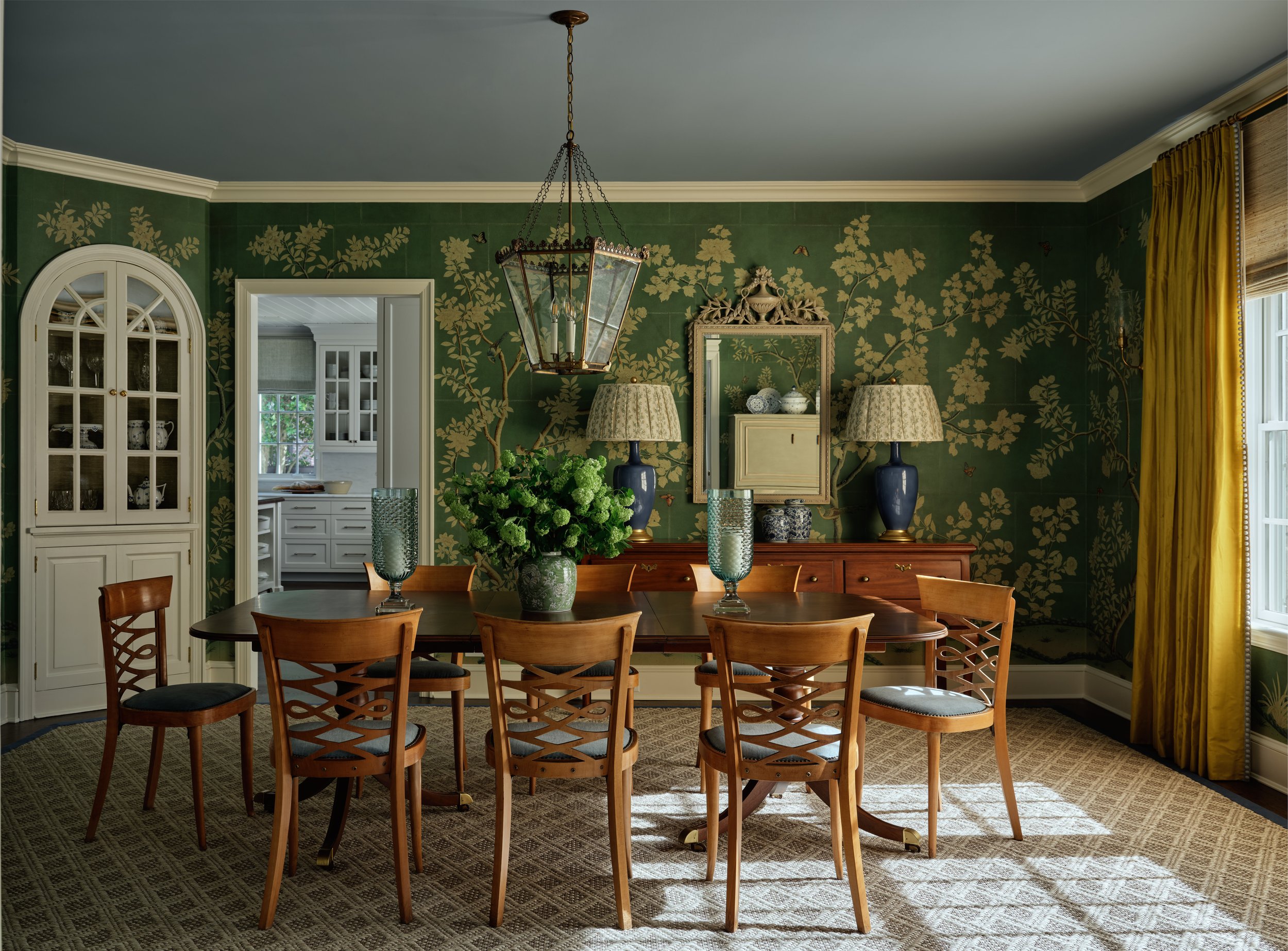 Dining room with dark wood table, eight chairs, a floral centerpiece, and a sideboard with lamps and decor, green floral wallpaper, and yellow curtains.