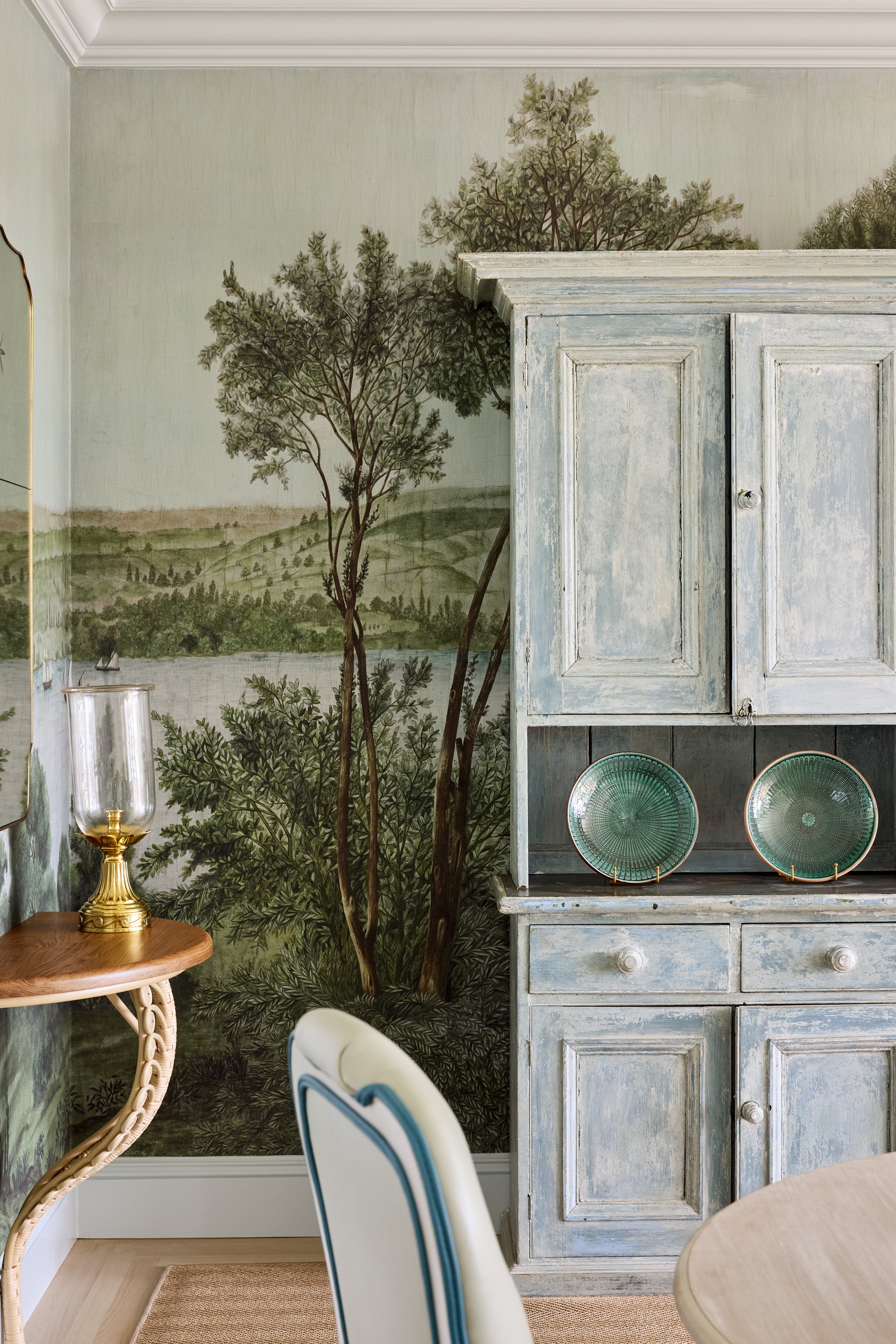 Decorative interior with painted landscape mural, distressed blue cabinet, two green plates on display, a small round wooden table with a brass oil lamp, and a white and blue chair.
