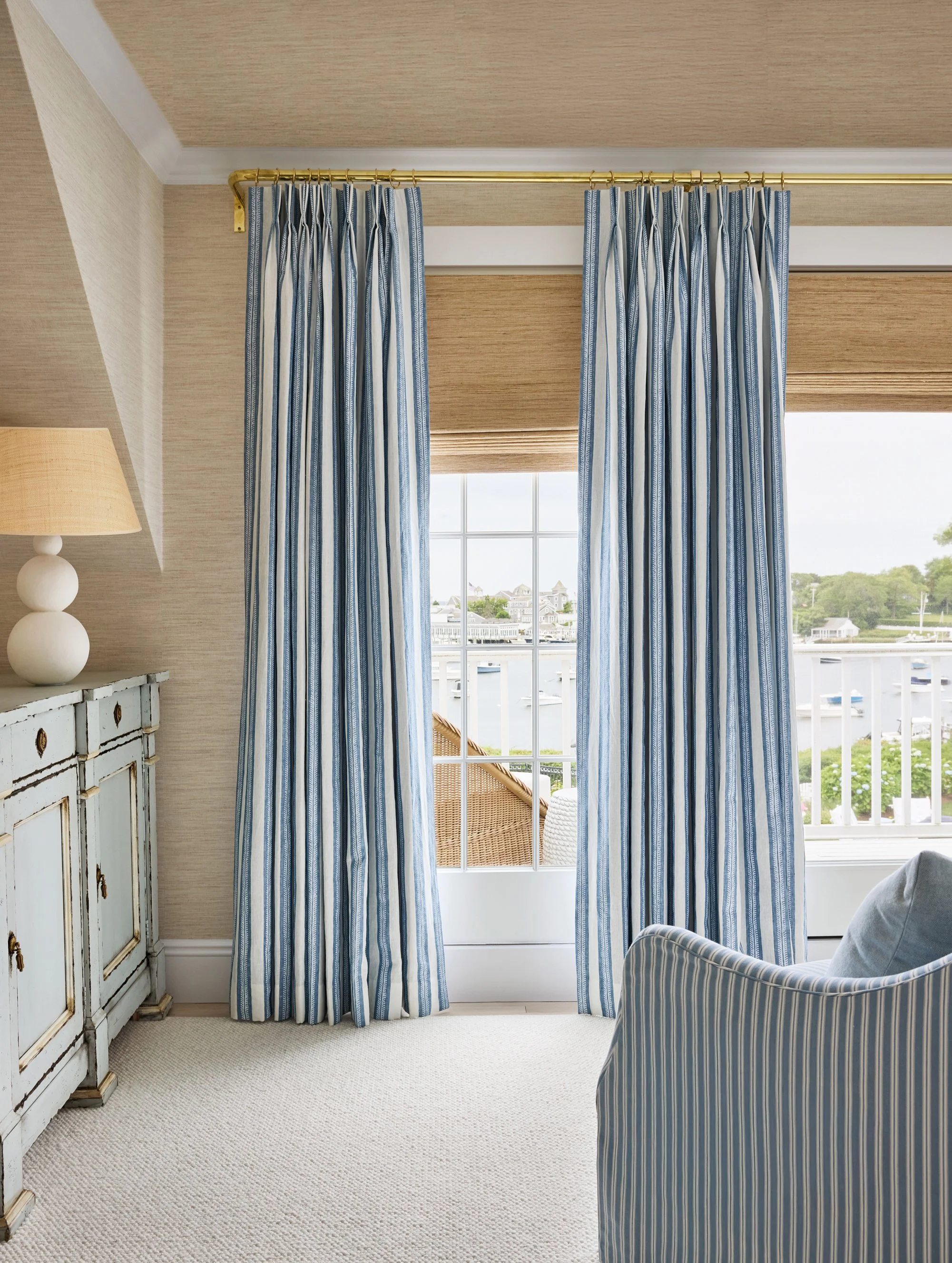 Room with blue and white striped curtains, a white wooden sideboard, a lamp with a round base, a striped armchair, and a window overlooking a marina with boats and houses.