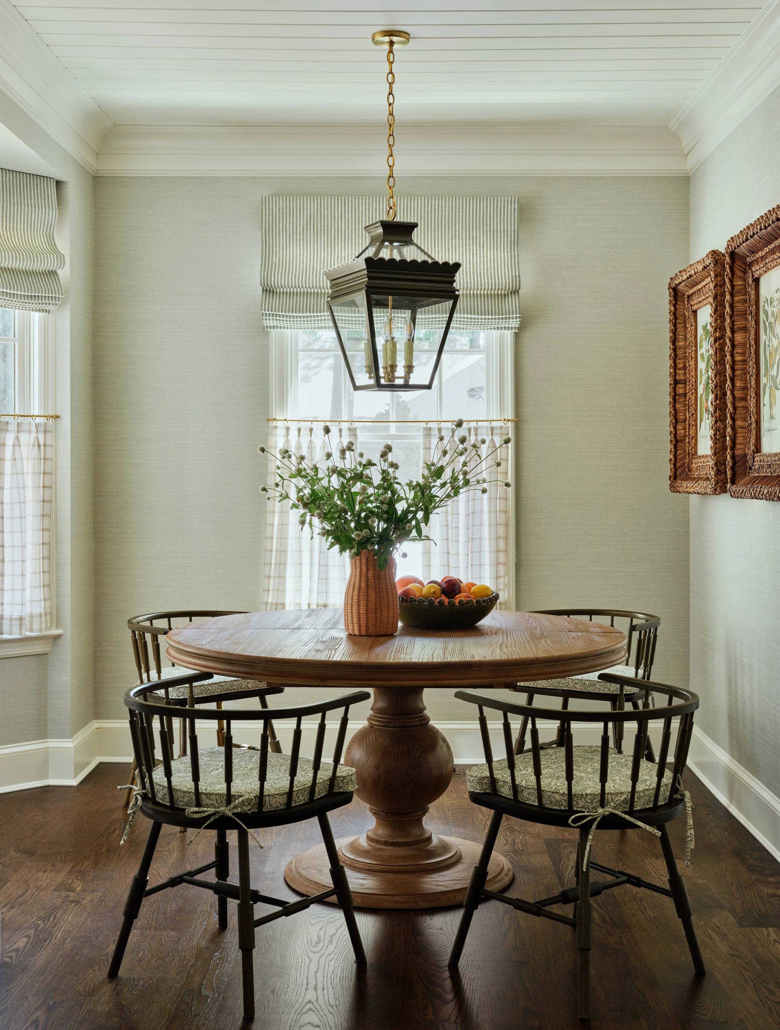 A dining room with a wooden round table, four black chairs with patterned cushions, a basket with flowers, a bowl of fruits, window with striped curtains, a hanging light fixture, and framed artwork on the wall.