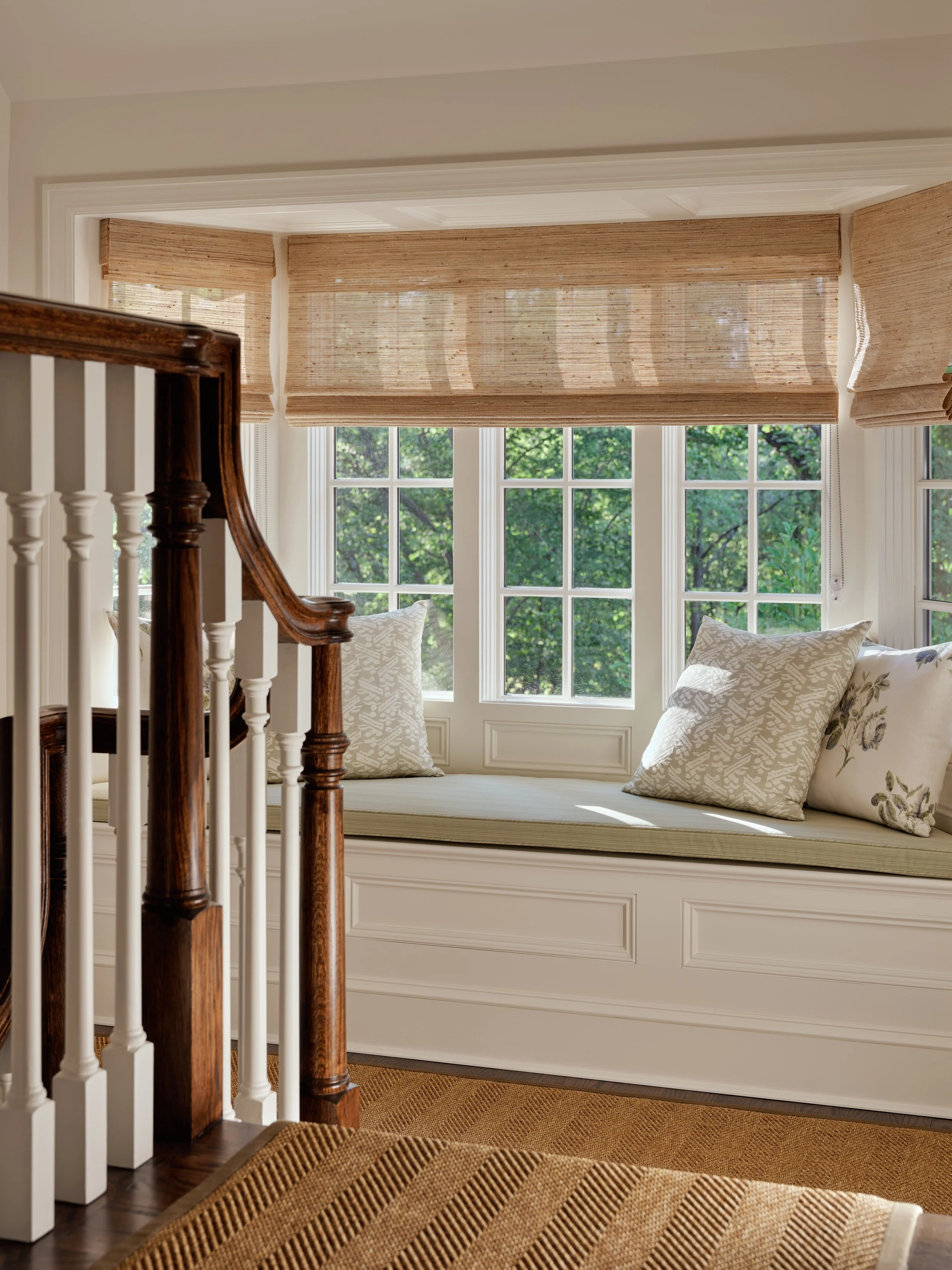 Sunlit window seat with beige cushions and pillows, framed by bay windows with bamboo blinds, in a cozy corner of a home.
