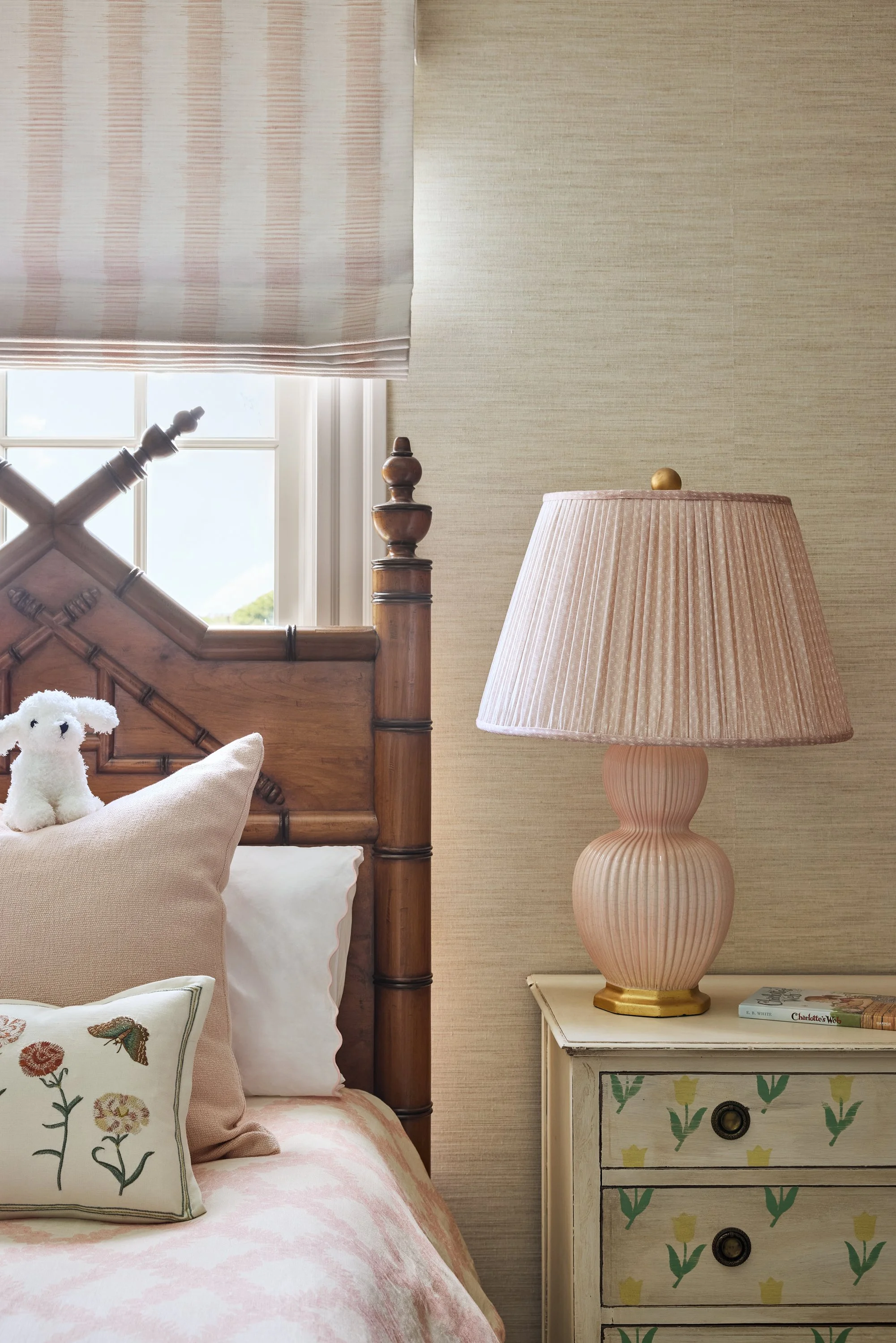 Close-up of a bedroom corner with a wooden bedpost, a pink bedside table with floral and bird pattern, pink and white bedding, a pink lamp on the bedside table, and beige wallpaper.