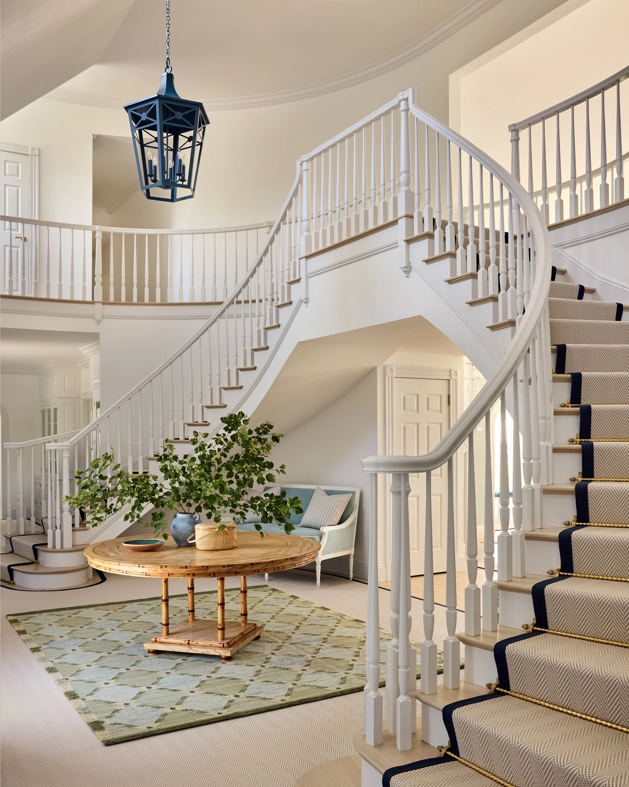 Interior view of a spacious, well-lit living area with a curved staircase, a wooden coffee table, a patterned rug, a green plant in a vase, and a light-colored bench with cushions. A blue chandelier hangs from the ceiling.