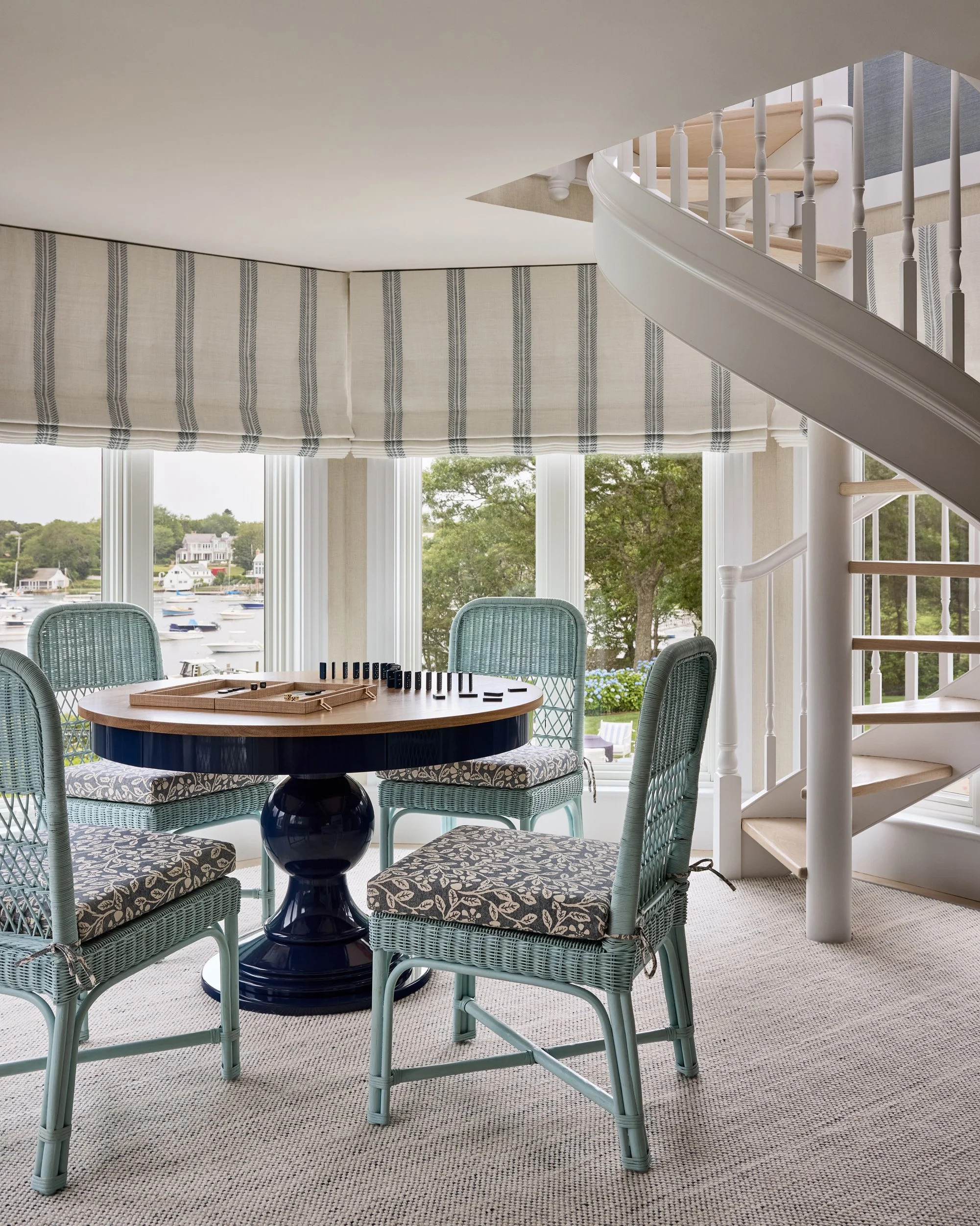 Interior of a house with a round table, four wicker chairs with patterned cushions, large windows overlooking a water view, and a staircase with white railing.
